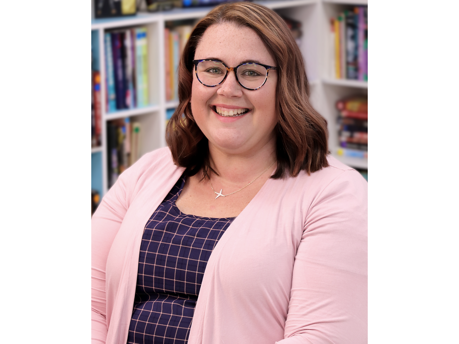 A woman with shoulder-length brown hair and glasses, wearing a pink sweater and necklace with airplane charm.