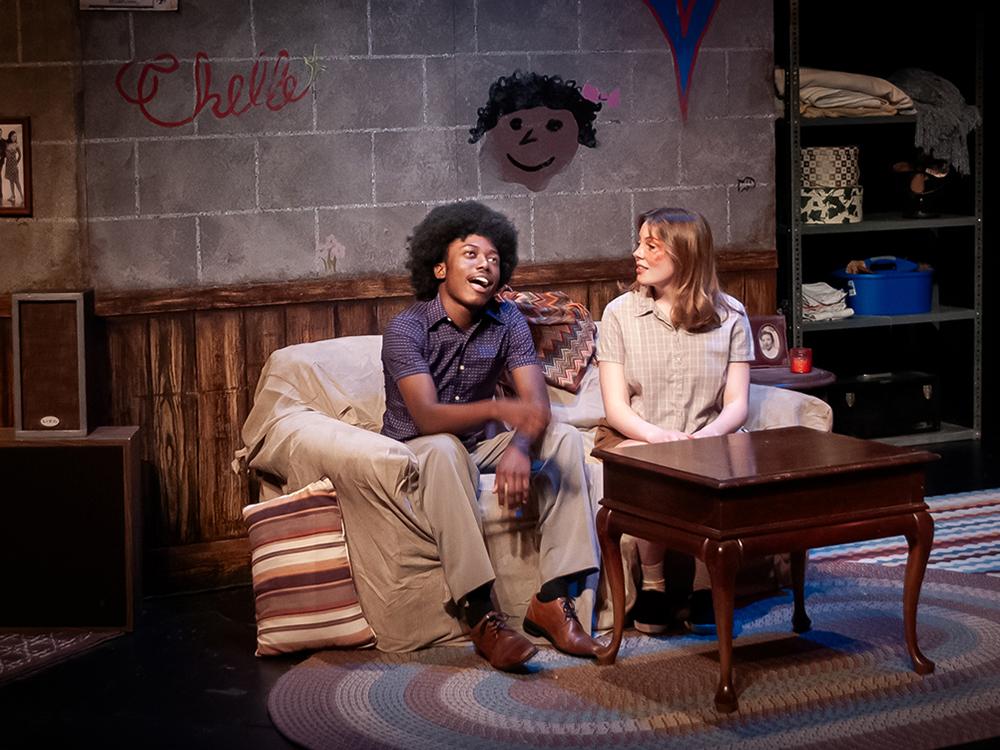 Two actors sit on a couch in an apartment decorated to look like it is from the 1960s