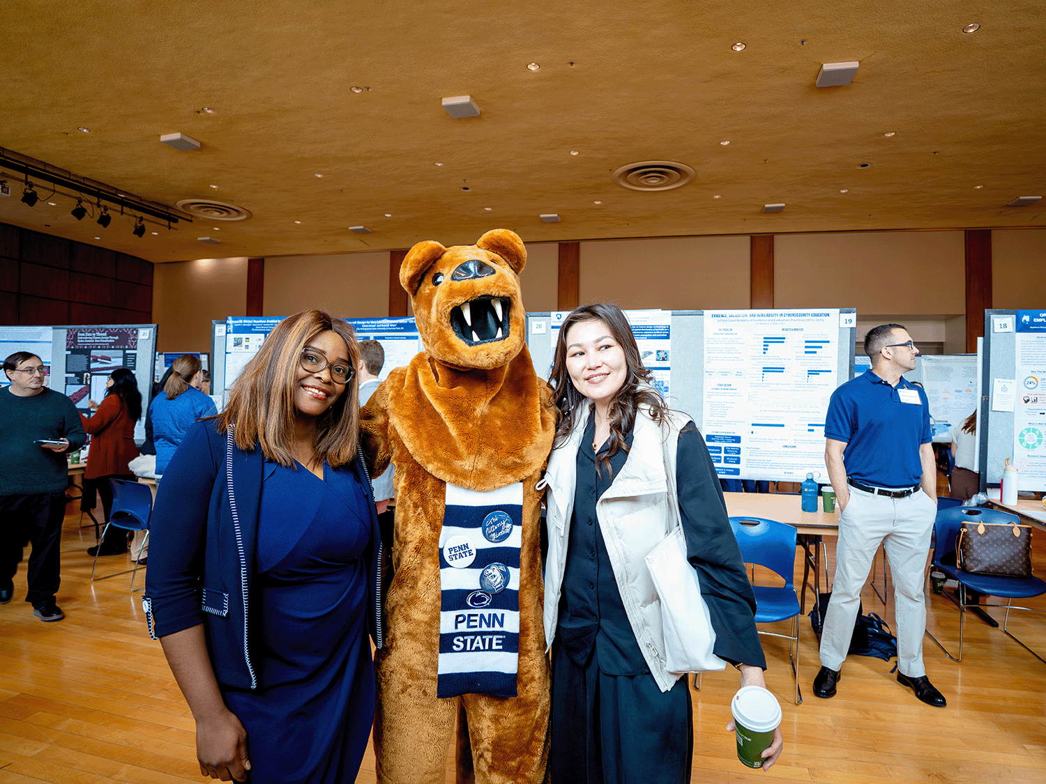 Nittany Lion mascot standing with two students at the 2026 Graduate Exhibition poster session