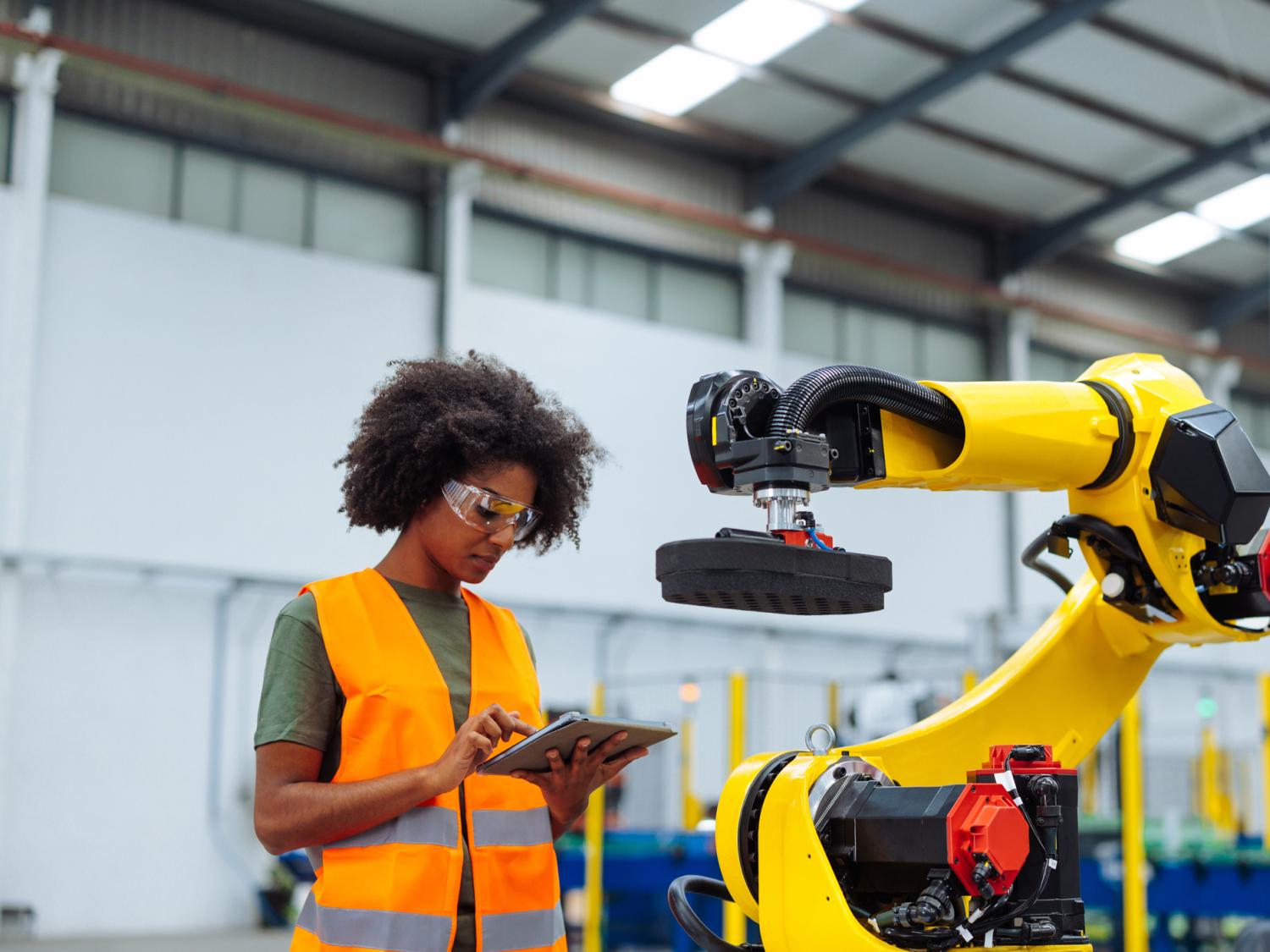 A woman wearing an orange safety vest uses a tablet to program an industrial robotic arm used in manufacturing