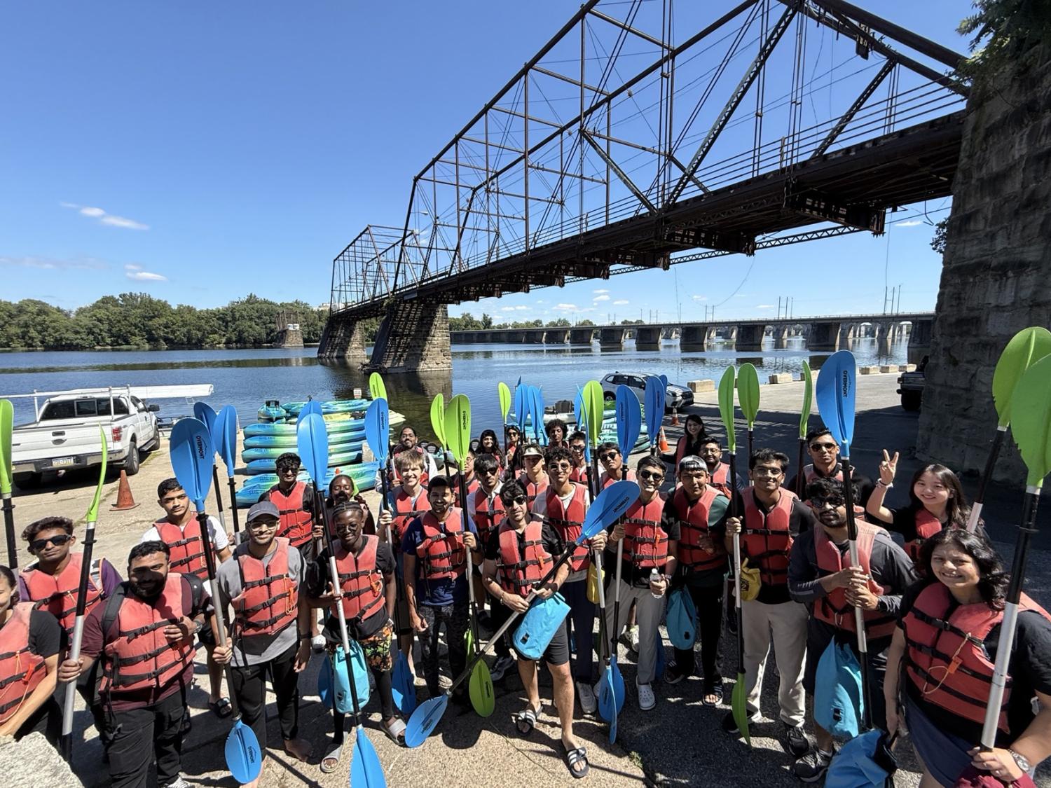 A group of people wearing life jackets and holding kayak paddles stand near a river