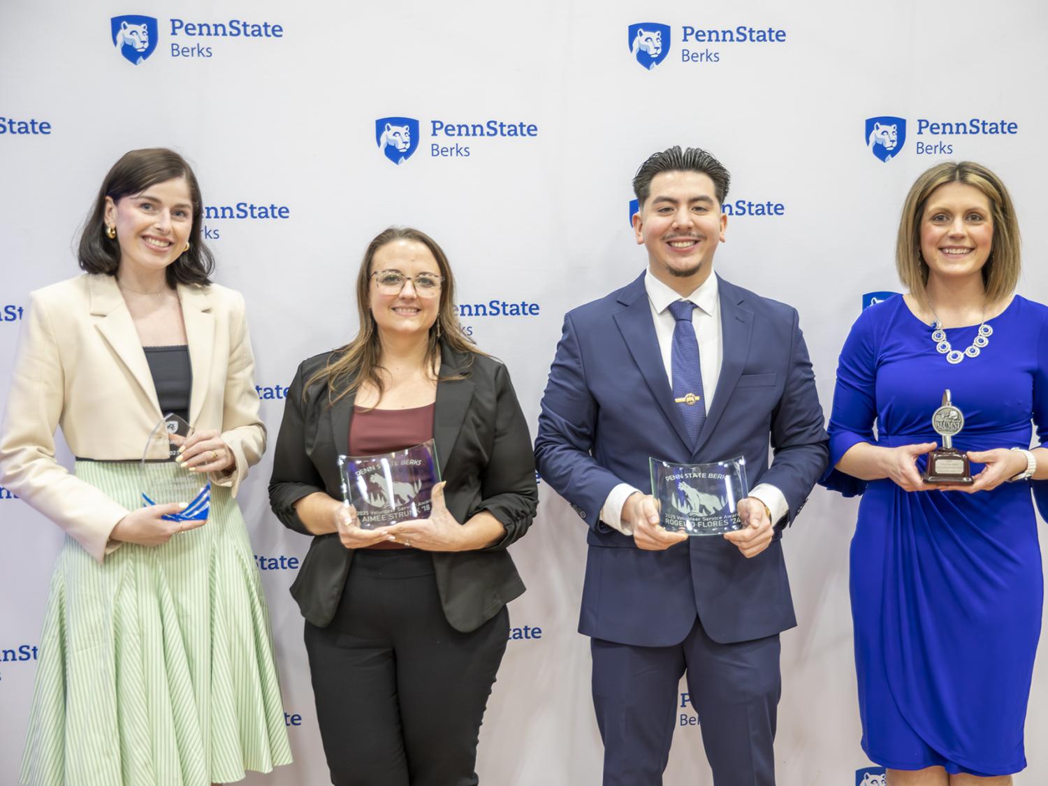 Four Penn State Berks alumni hold awards and stand in front of step-and-repeat logo banner