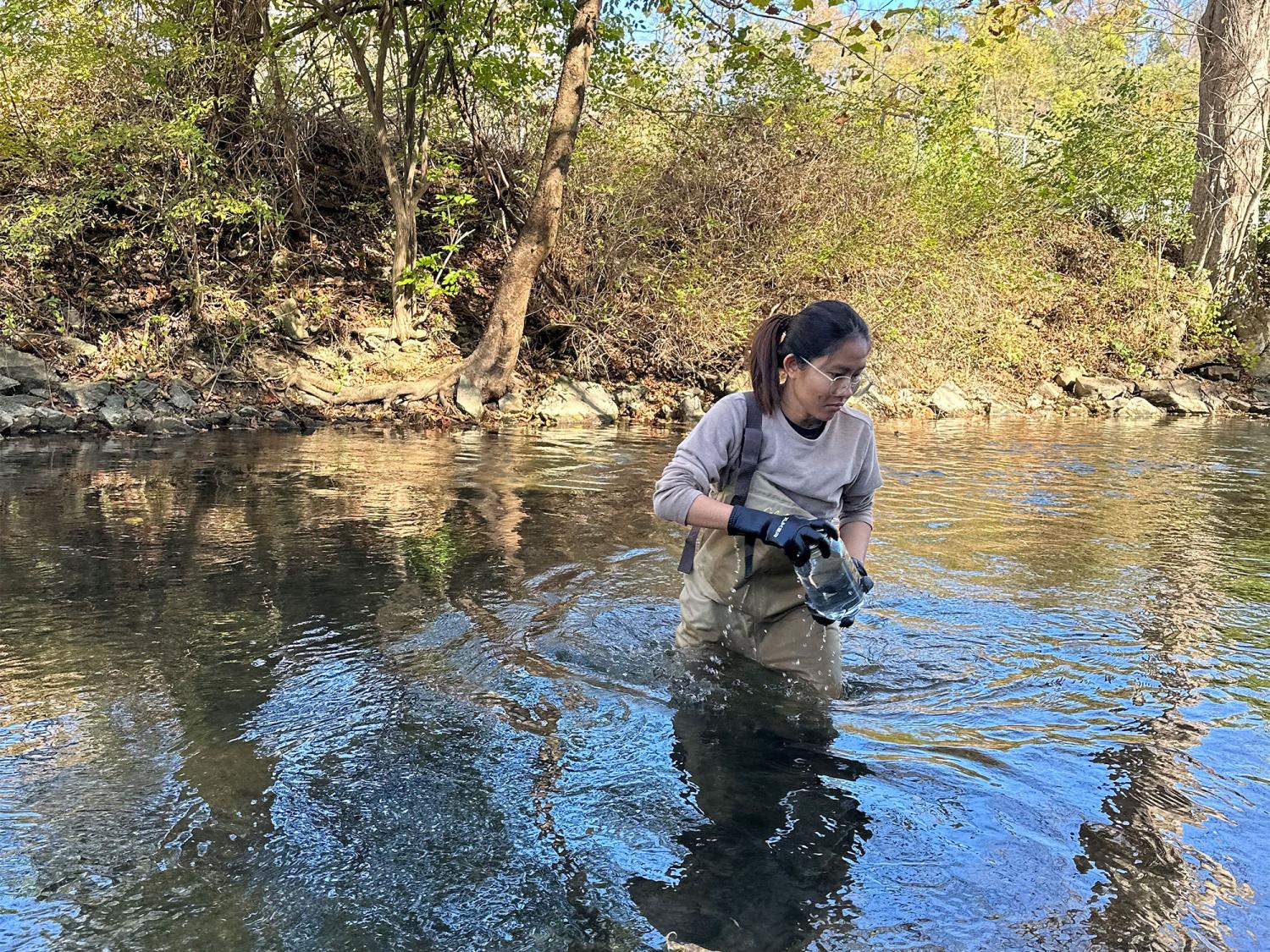 A researcher, waist deep in water, collects water and sediment samples from a river in a glass jar