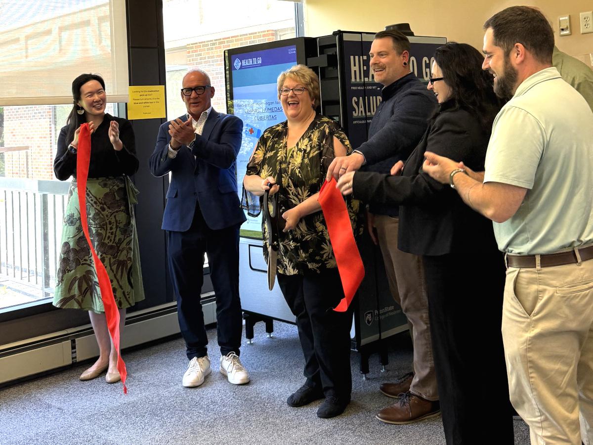 Six people smile and clap immediately after cutting a ribbon, as they stand in front of a vending machine. A window is also in the background.