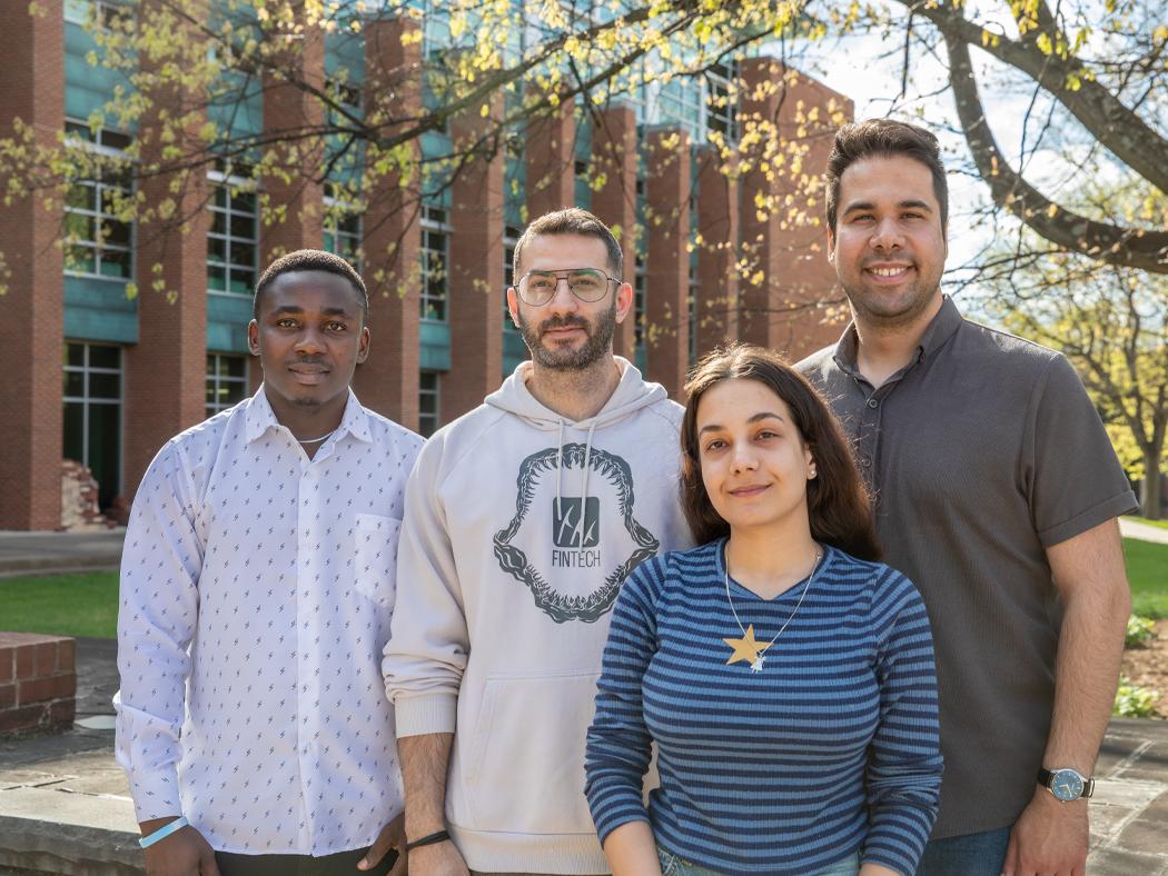 Four people stand together outdoors in front of a brick and glass campus building, with trees overhead. They are casually dressed, facing the camera, and positioned closely in a small group on a sunny day.