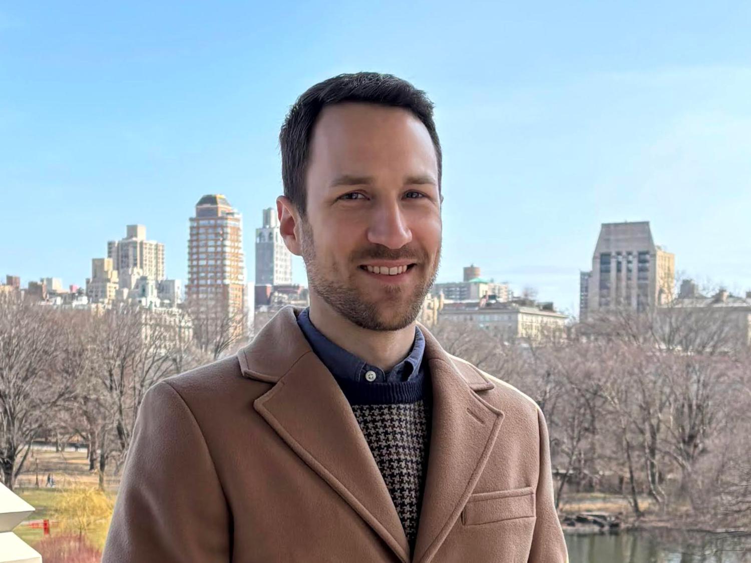person with short dark hair and beard in woold coat with city skyline in background