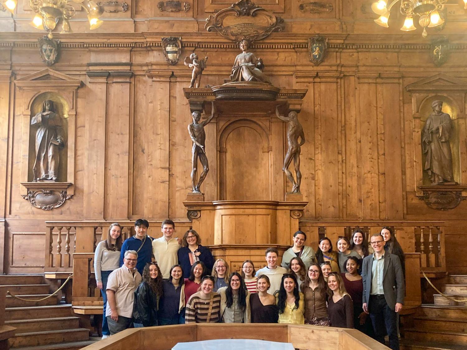 Students stand in front of Anatomical Theatre, brown space, with chandeliers, students taking up a few rows.