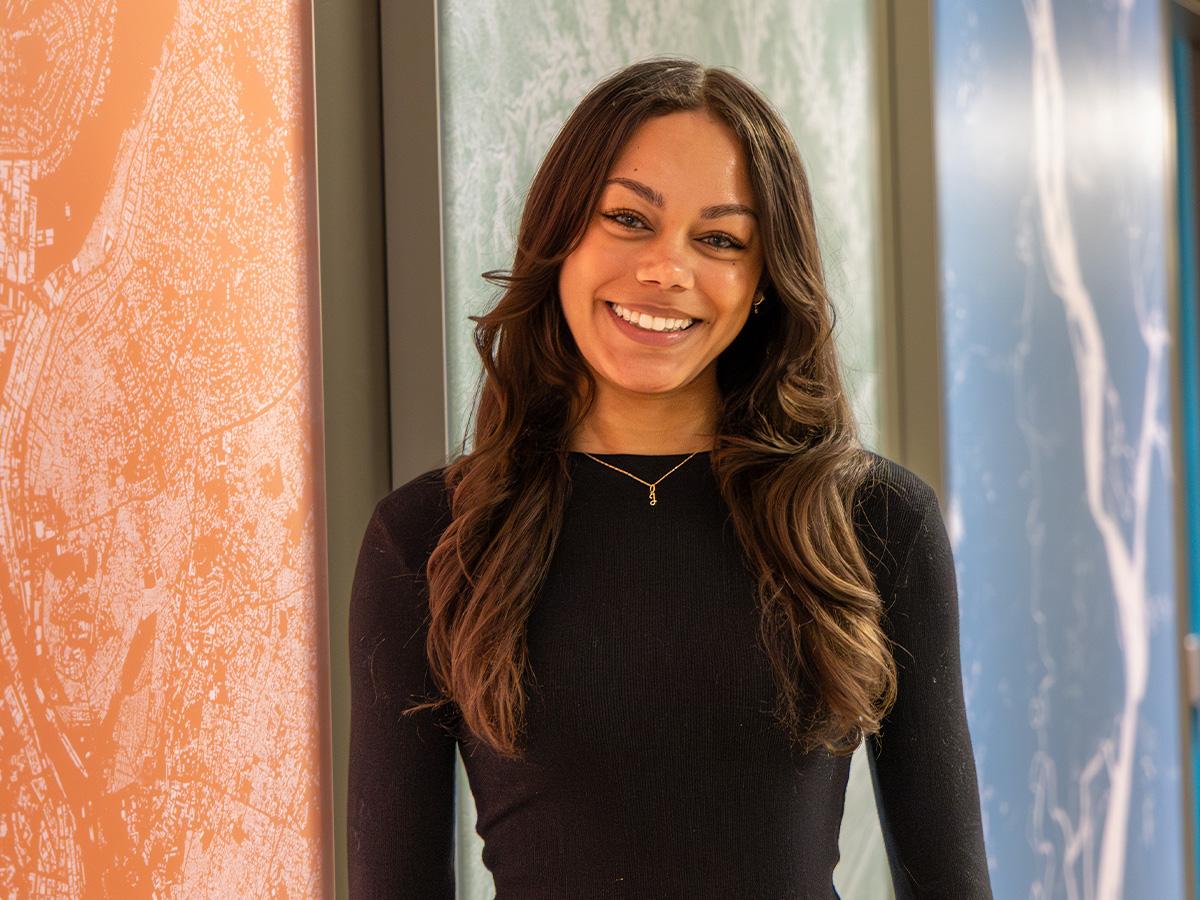 Jazmyn Clark wears a black top stands in front of a multicolored map panels that are orange, green, and blue hues.