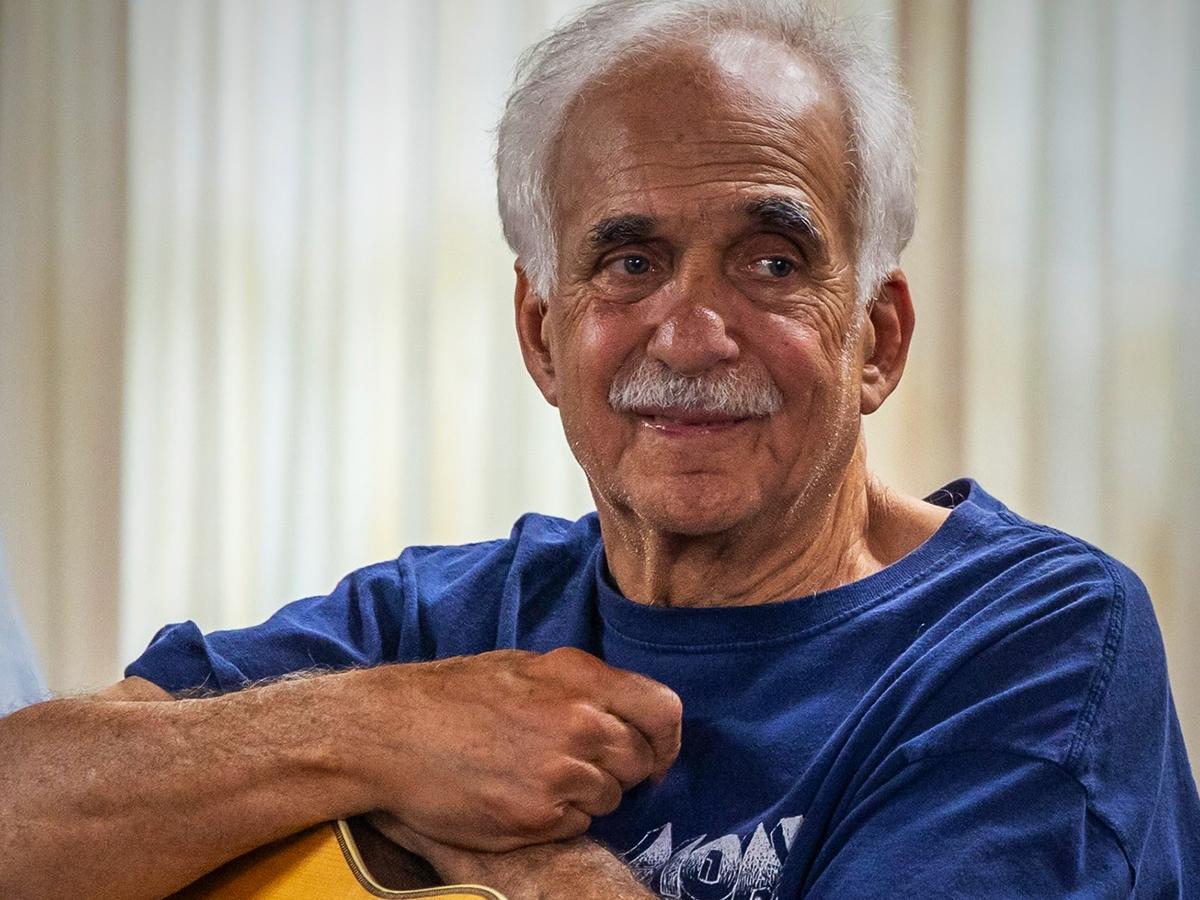 Jerry Zolten smiles while holding a guitar, seated indoors against a softly lit background.