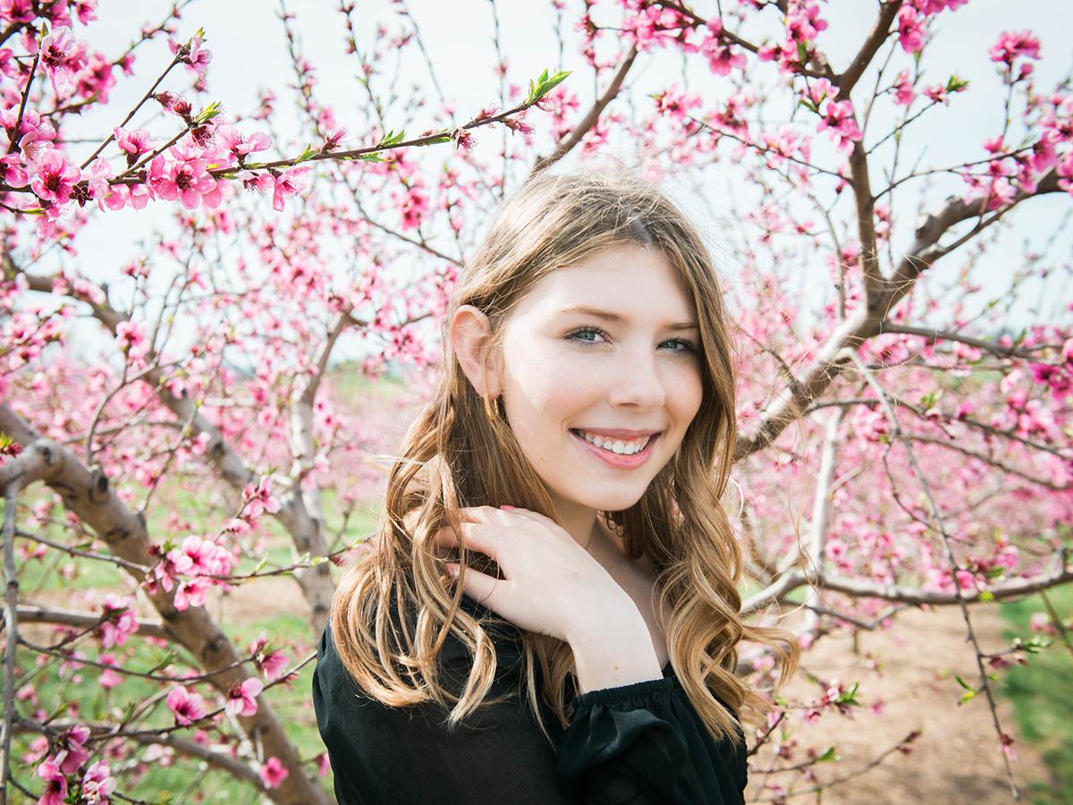 A teen girl in front of a flowering tree smiles at the camera. She has long hair and is touching her shoulder.