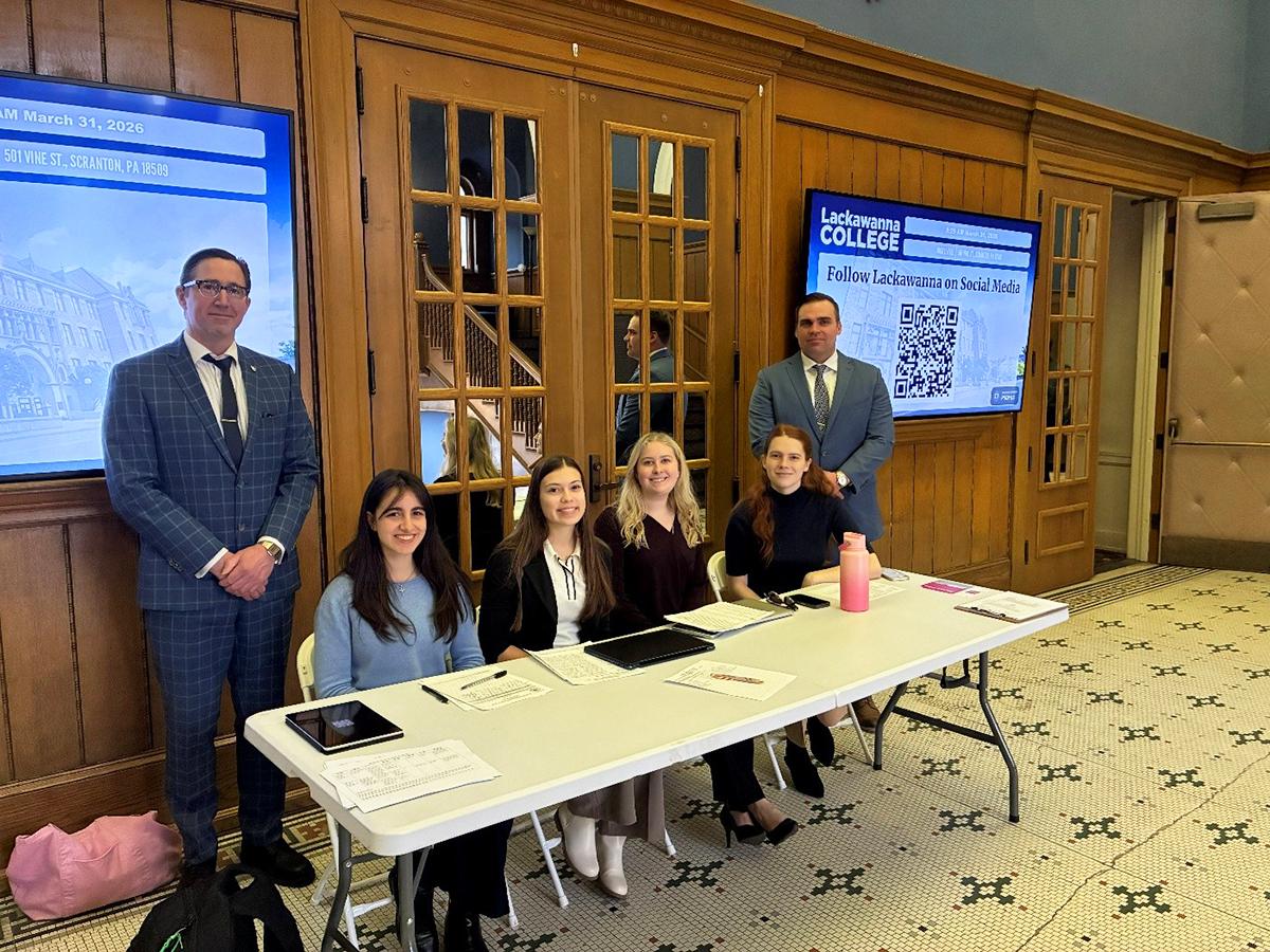 A group of students sit behind a registration table with materials and tablets, while two suited professors stand behind them, in a formal indoor venue with digital displays in the background.