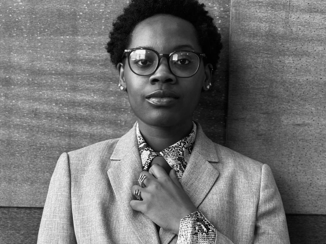 Black-and-white portrait of a person wearing round glasses and a light-colored suit jacket, standing against a textured wall.