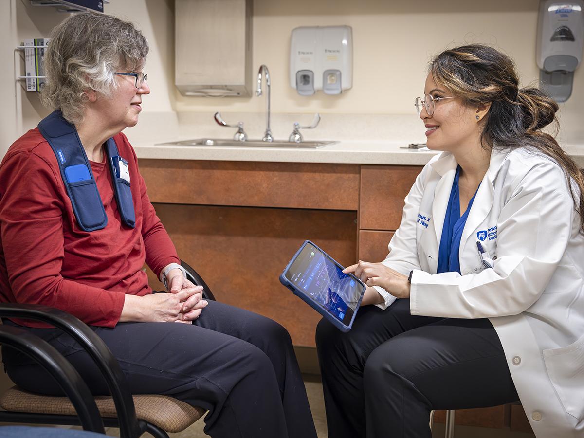A woman patient sits across from a neurologist in a medical office. The patient has an adaptive deep brain stimulation device around her neck. The neurologist touches a tablet to adjust the device and smiles at the patient. 