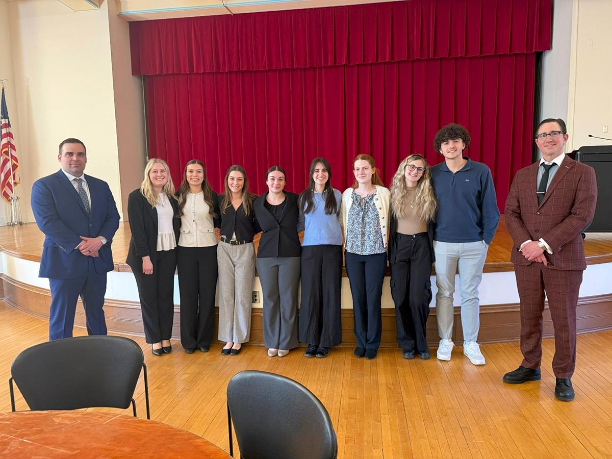 A group of students stand together on a stage in front of red curtains, flanked by two professors on either end, posing for a photo in a formal indoor setting.