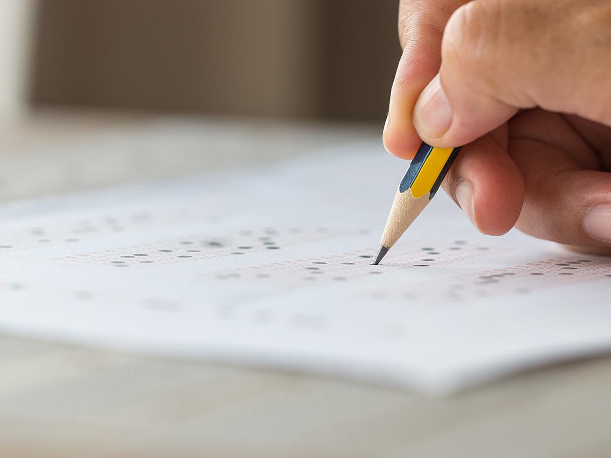 A close-up shows a hand filling in answers on a standardized test sheet with a sharpened pencil.