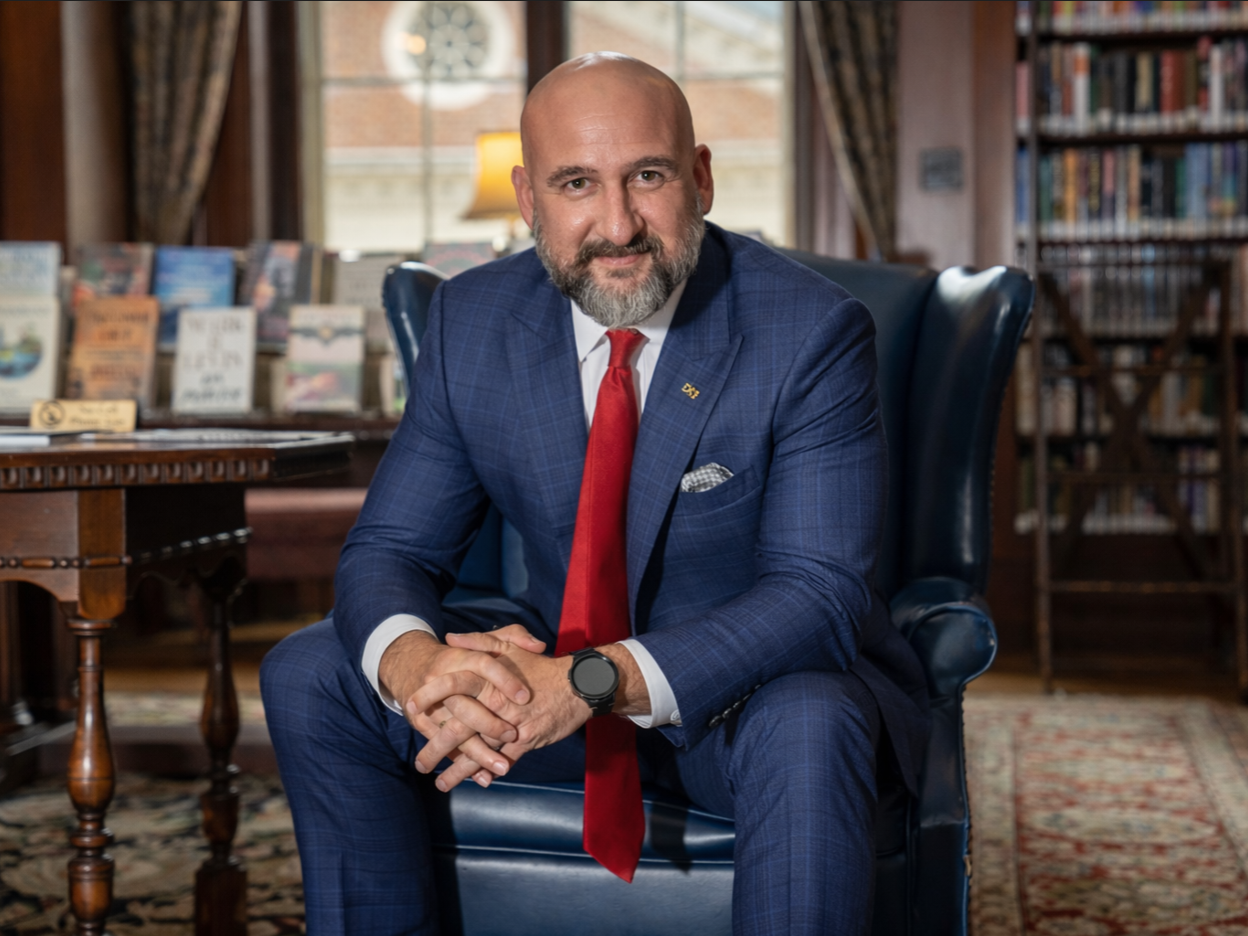 A man in a business suit sits in a library, learning forward and smiling.