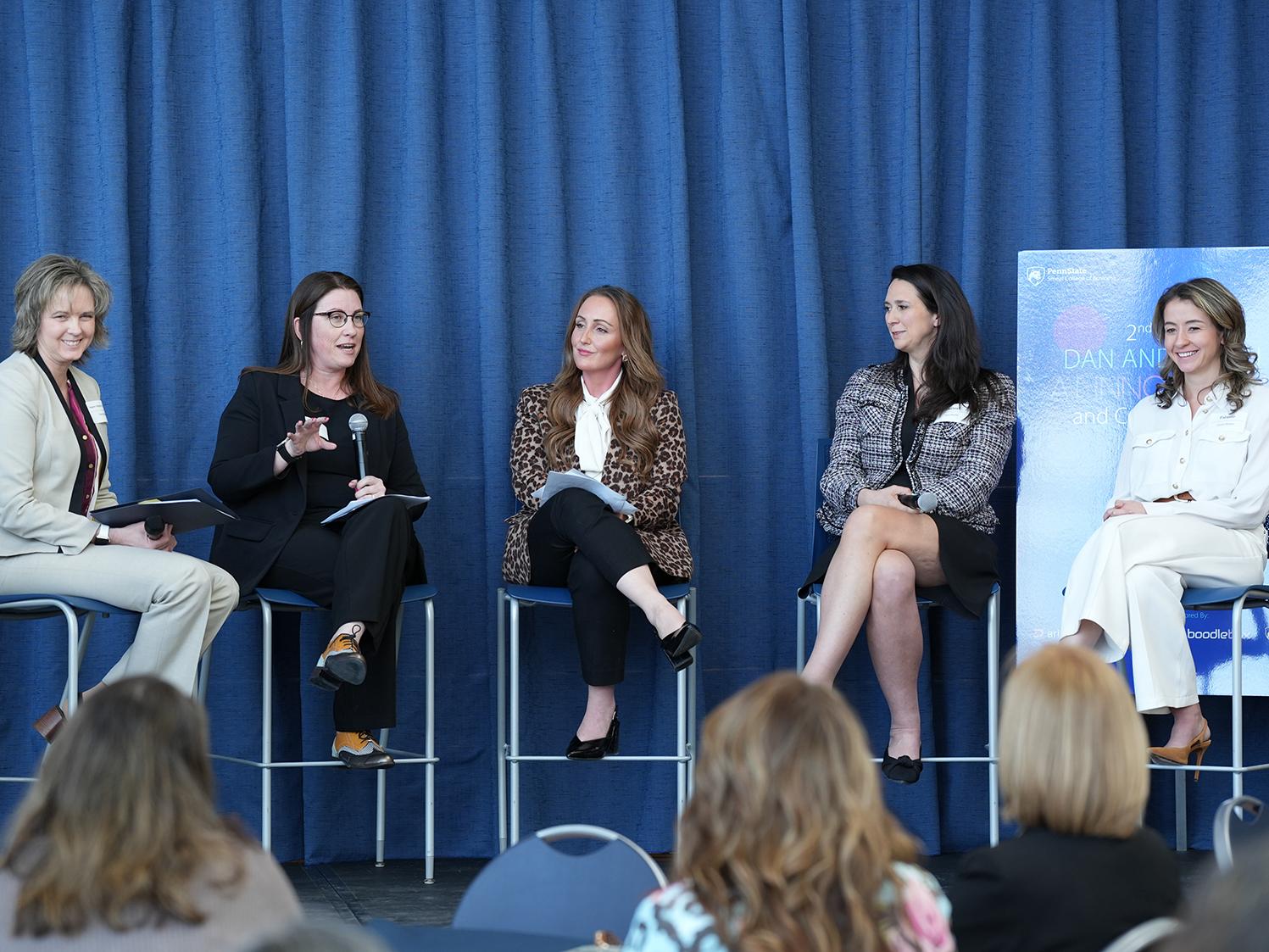 Five women sit on stools on a stage. One holds a microphone.