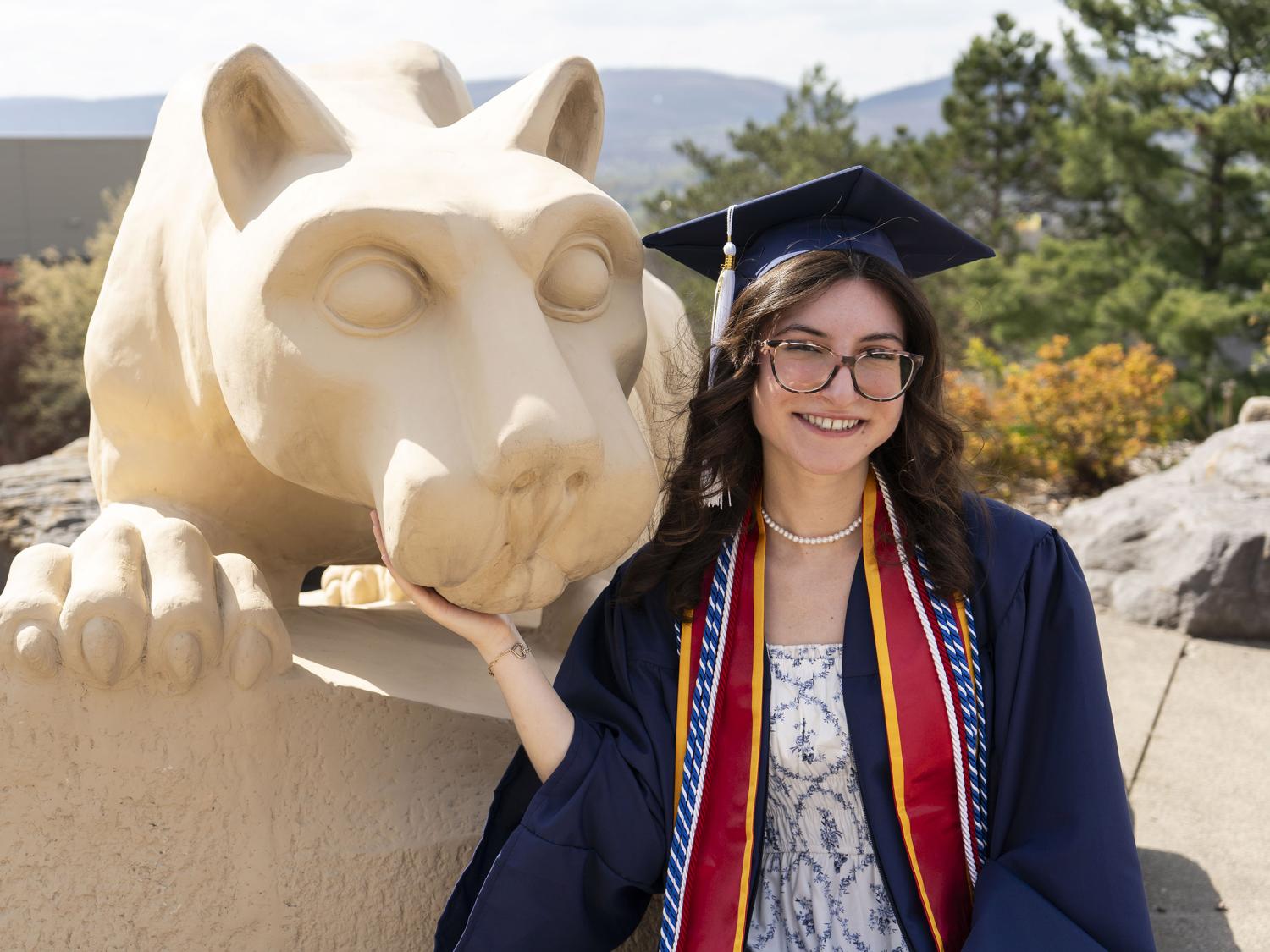 Female graduate, wearing commencement regalia, smiles next to Lion Shrine at Penn State Scranton