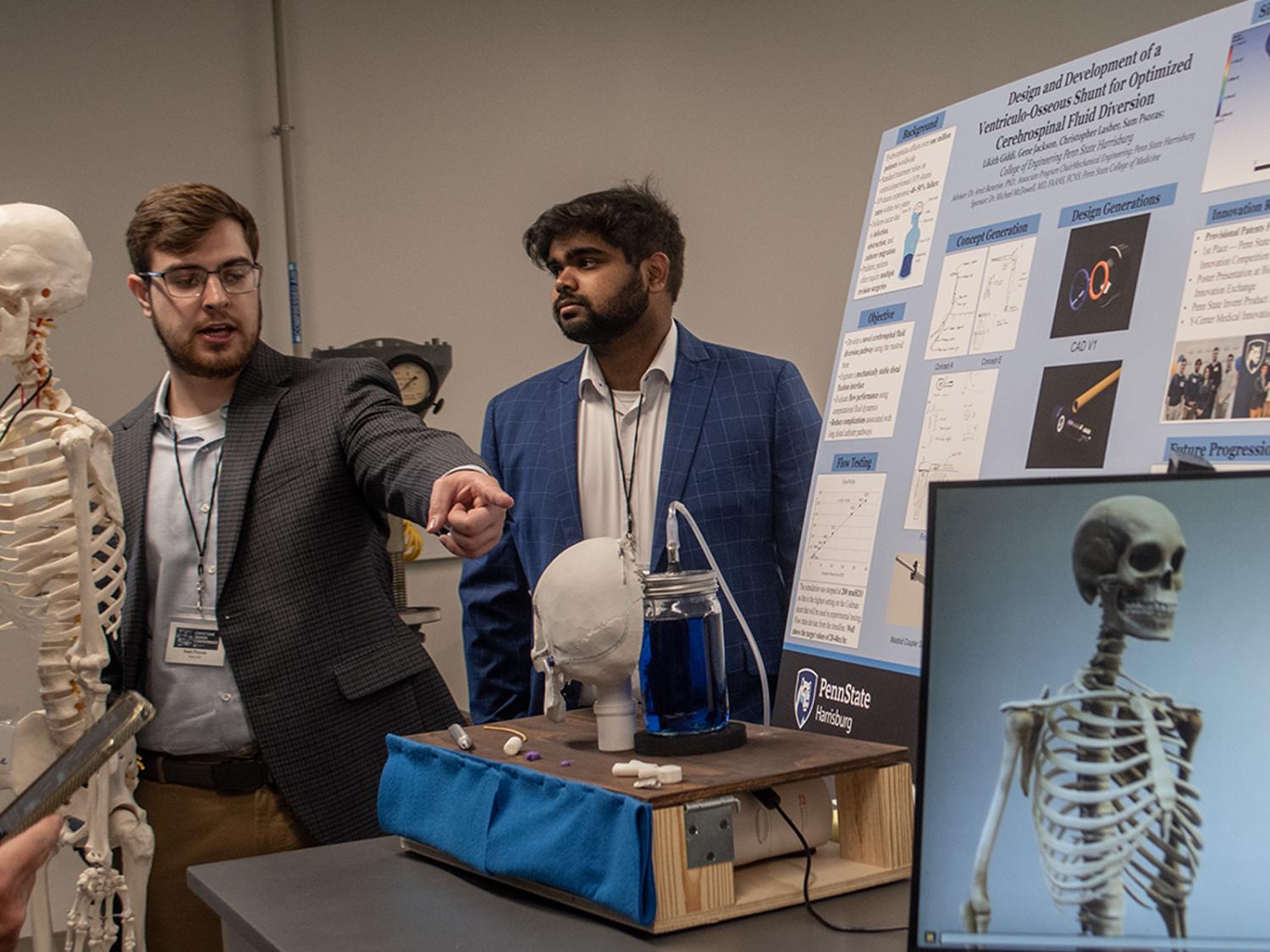 Two students, standing near a poster and a skeleton, explain their capstone project to guests