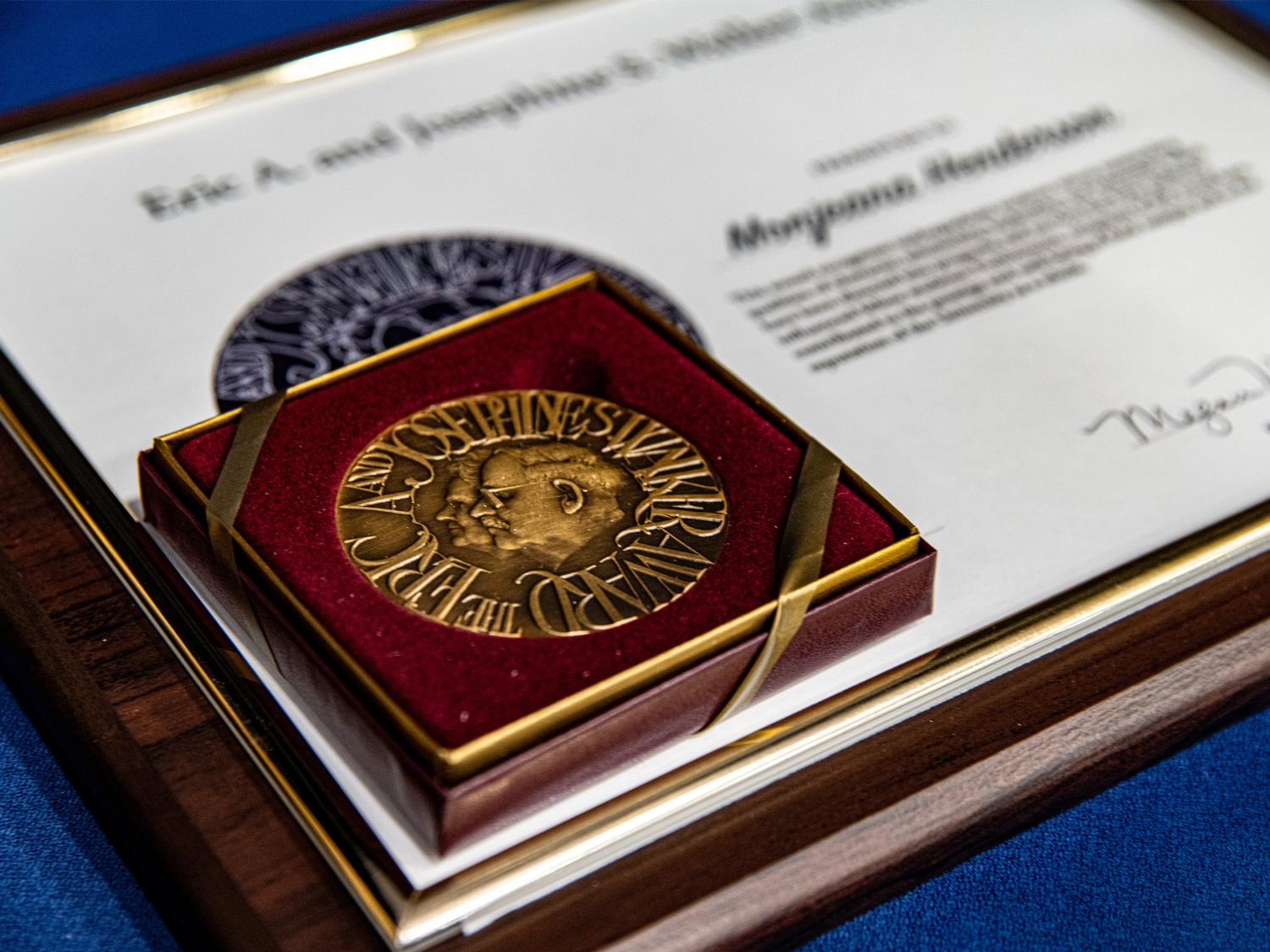 A bronze medal in a red velvet presentation box rests on top of a framed certificate on a table covered with a blue cloth. The certificate includes the name Monjeana Henderson and formal text recognizing an award.