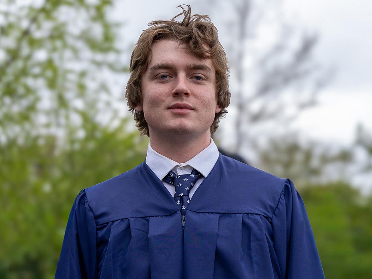 Sailor Walter in a blue graduation gown and tie stands outdoors. The background features blurred green foliage and a cloudy sky.