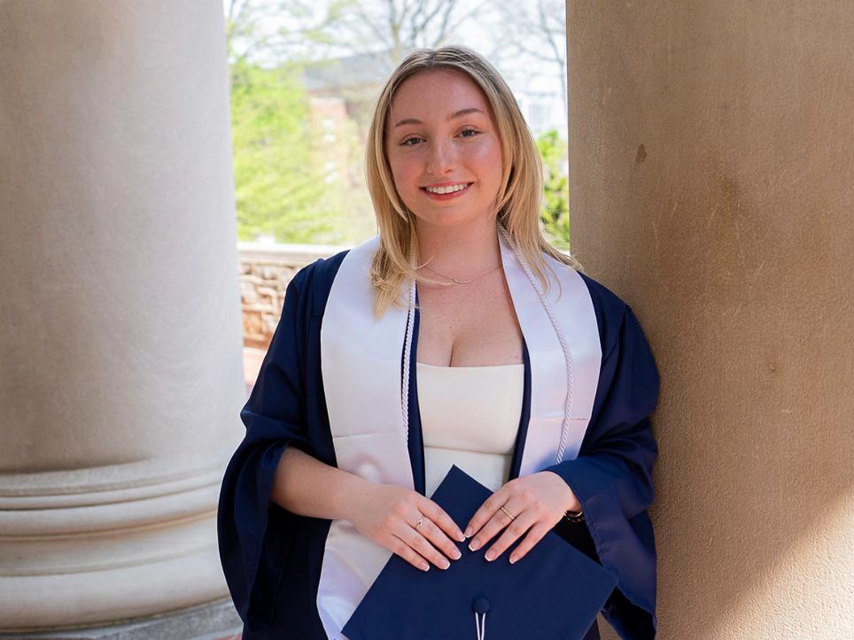 Abby Fusaro stands in a graduation gown against a pillar outside Old Main Building.