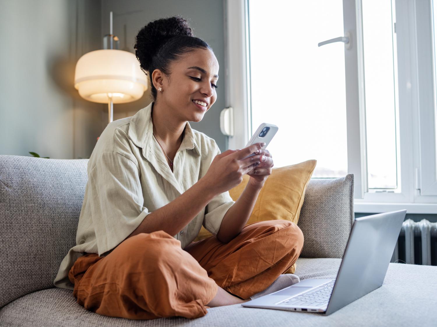 A person sitting cross-legged on a couch, smiling and looking at a phone. A laptop is also on the couch.