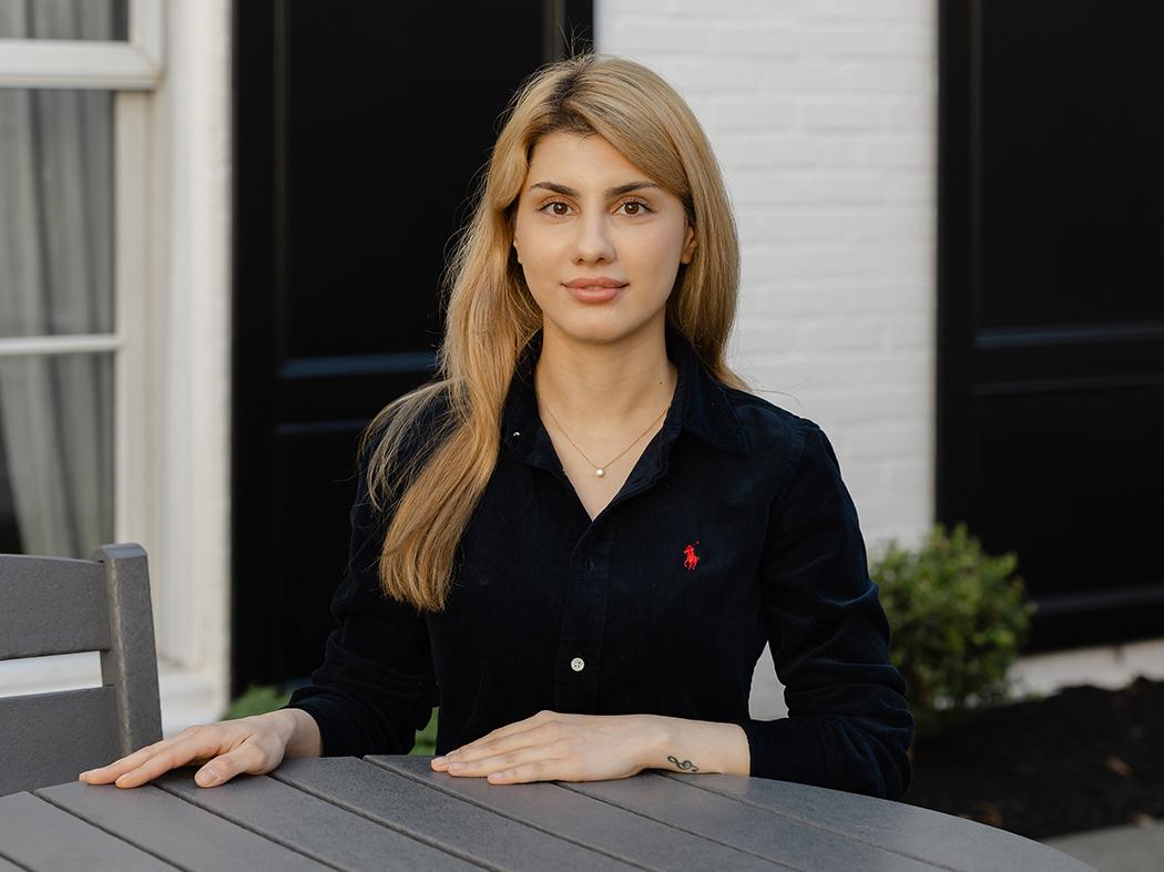 A person sits at an outdoor table, facing the camera with hands resting on the tabletop. The person has long, light-colored hair and wears a black button-up shirt with a small red embroidered logo on the chest and a delicate necklace. 