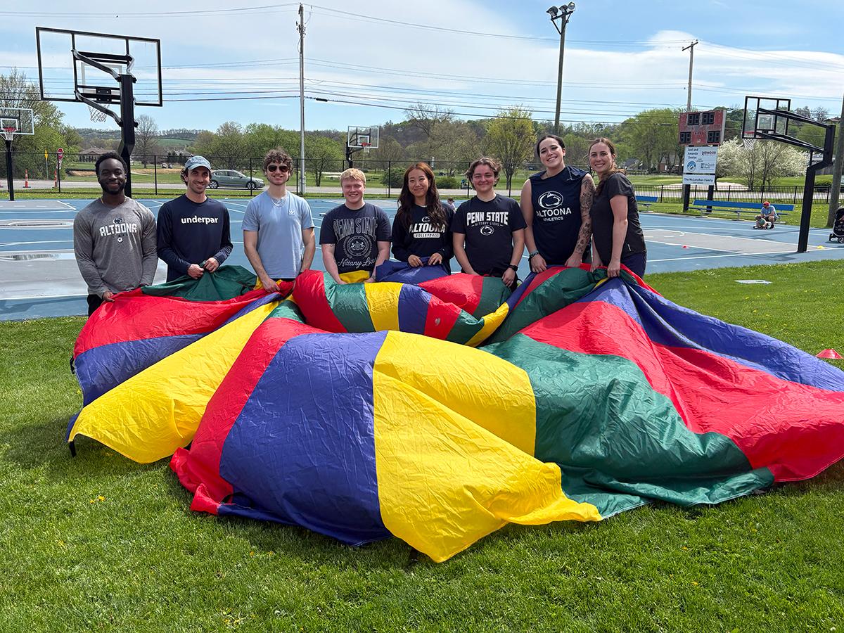 Group of students stand on grass holding a colorful parachute near outdoor basketball courts on a sunny day