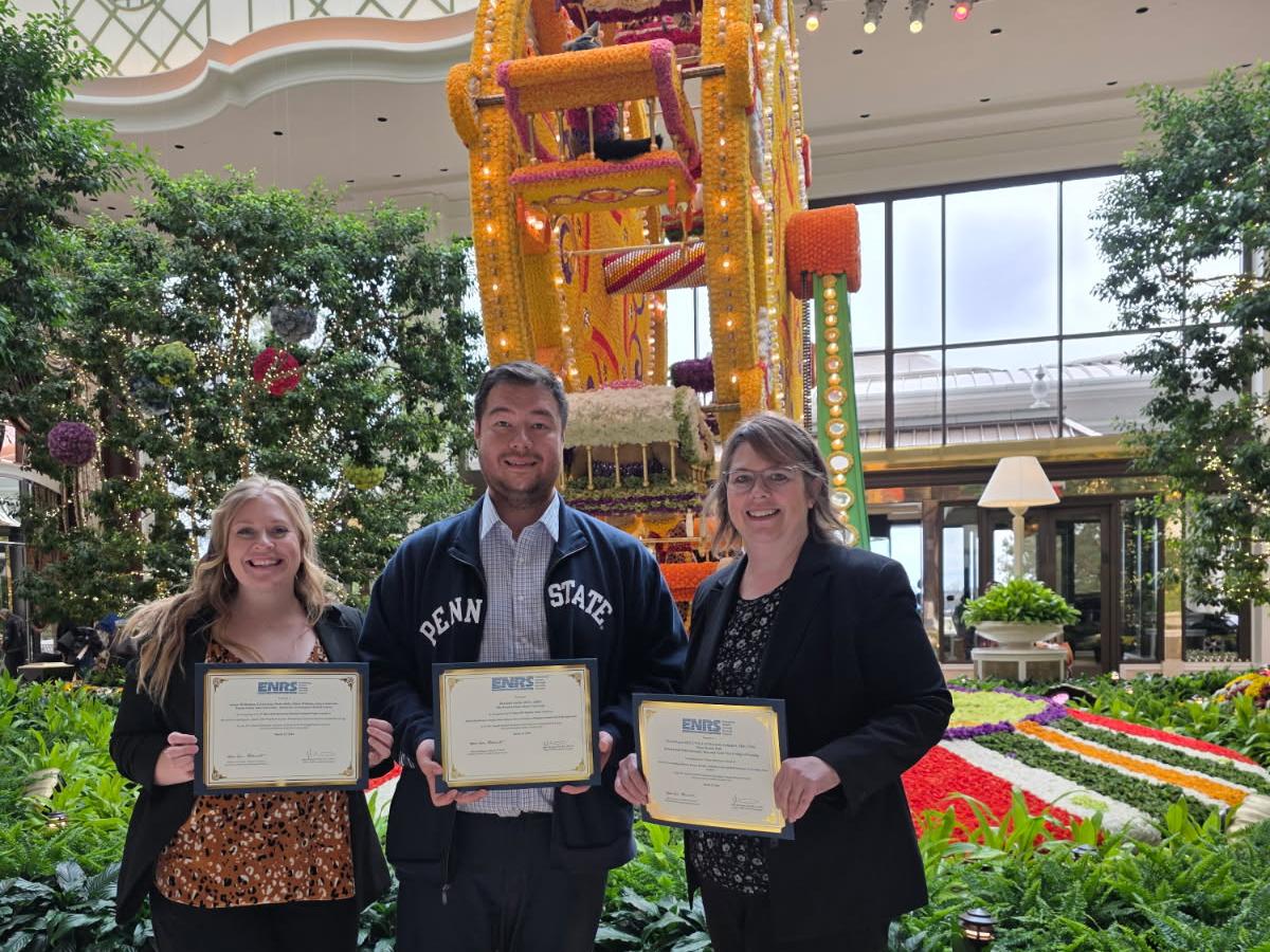 Three people standing indoors holding certificates with a large colorful floral Ferris wheel sculpture and lush greenery in the background.