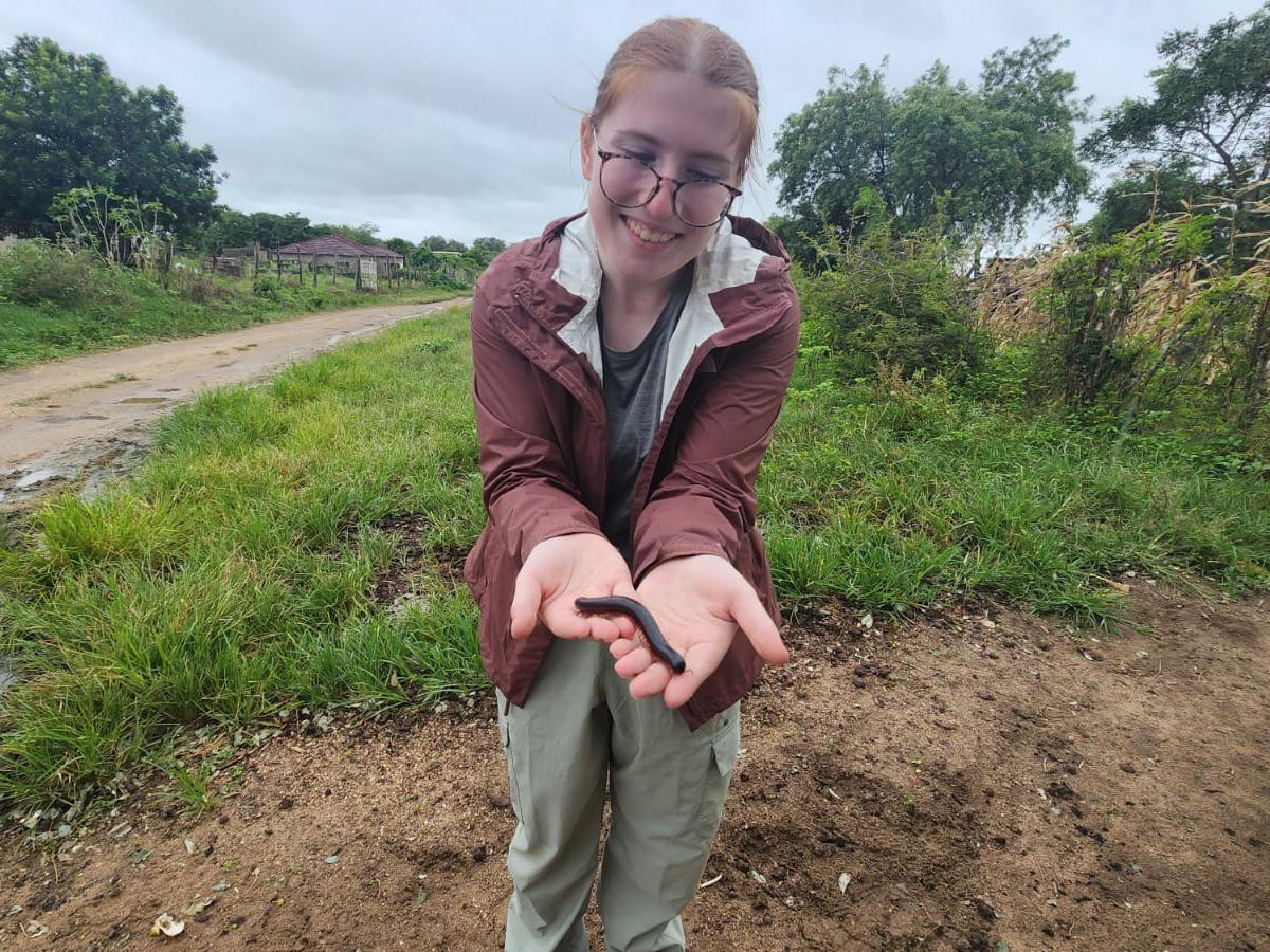 Isabel Rivera wearing glasses and a brown jacket stands on a dirt path, holding a small black creature in their hands and smiling. Greenery and cloudy sky are in the background.