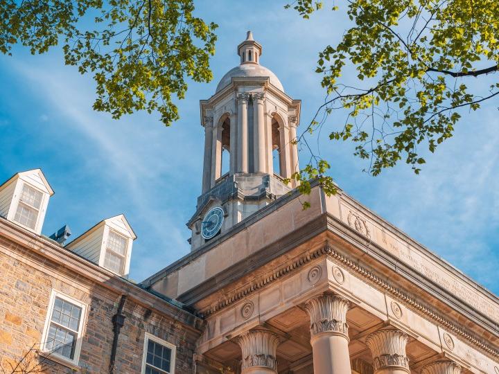 A ground-up shot of the Old Main bell tower with green leaves in the foreground and a blue sky in the background