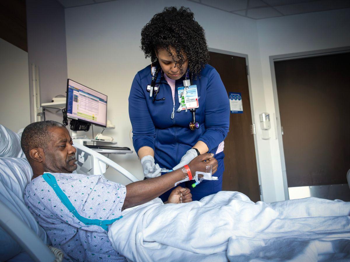 A nurse in a hospital room adjusts an IV line on a male patient lying in a bed, while monitoring equipment and a computer are visible in the background.