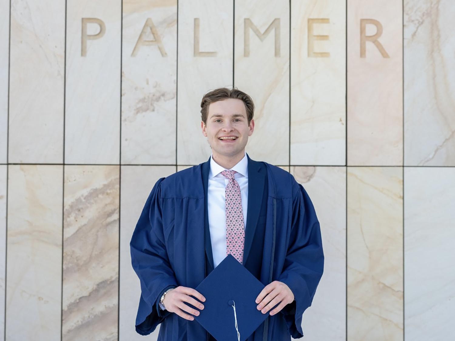 A photo of a smiling Palmer Moore in a commencement gown, holding a graduation cap and standing in front of the words PALMER on a wall behind him.