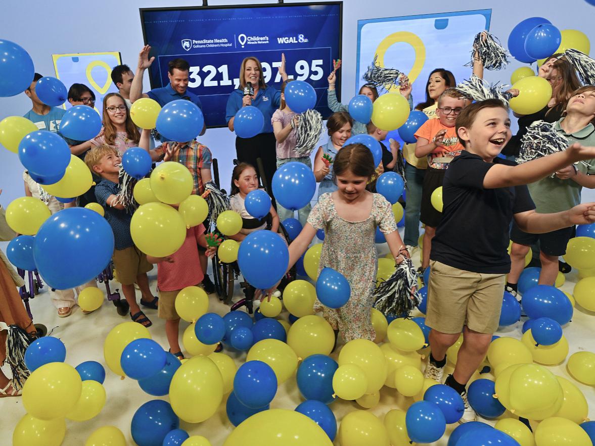 Children and event hosts celebrate during a Children’s Miracle Network fundraising reveal, tossing blue and yellow balloons into the air in front of a large display showing the total raised. The room is filled with smiling children, balloons, and pom-poms as participants cheer during the announcement.