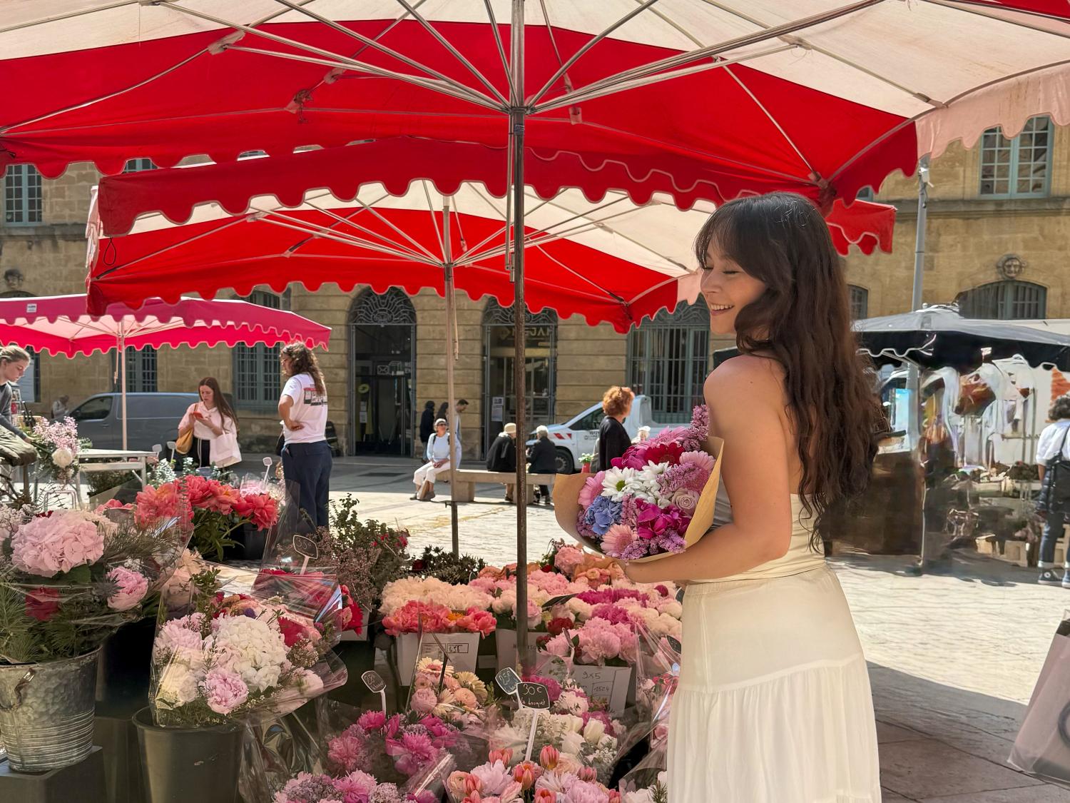 Jenna Guarrera wears a white dress while smiling and holding a bouquet of pink, purple, and white flowers in front of a flower stand on the streets of France.