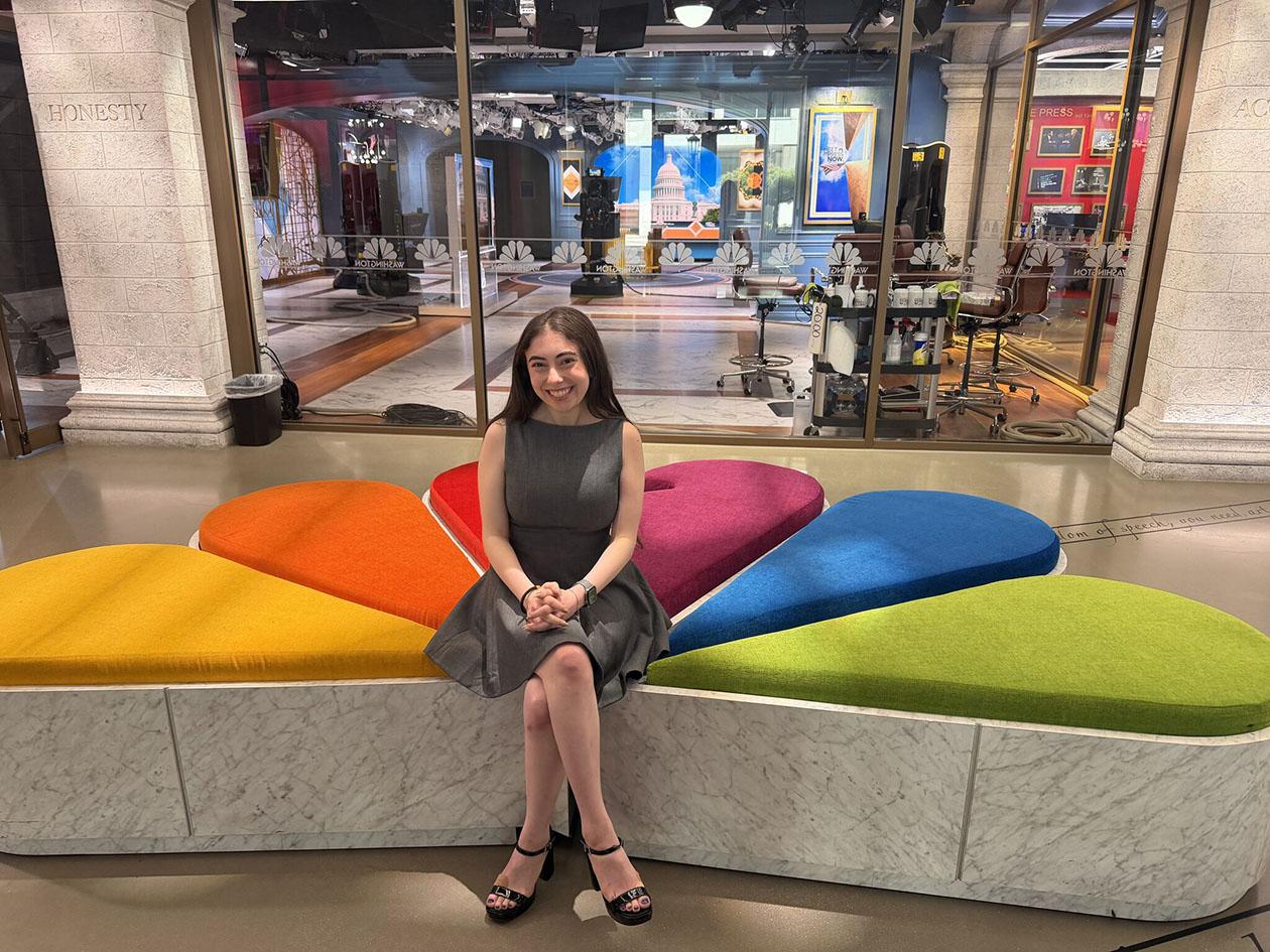 A dark haired woman sits on a multicolored couch shaped like a peacock silhouette.