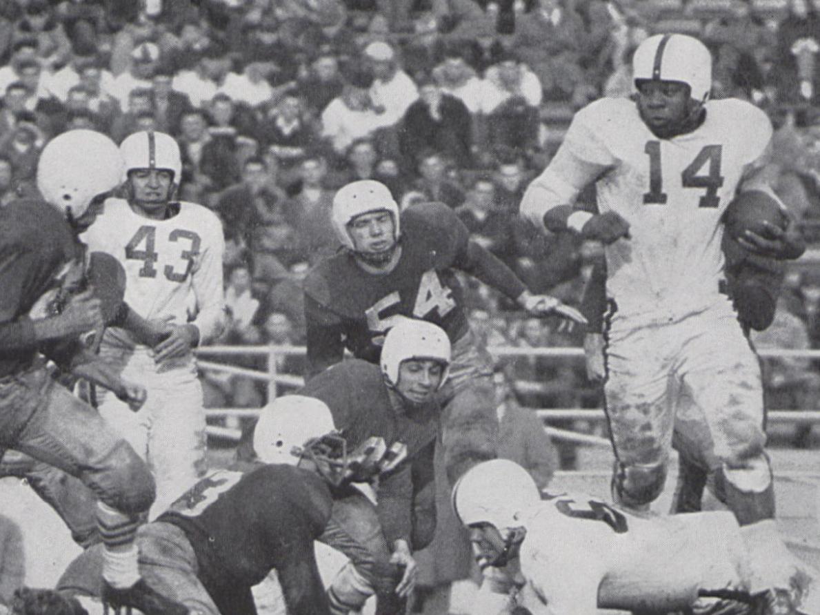 Several football players in dated uniforms are captured in mid-play while a crowd sits in the bleachers in the background. 