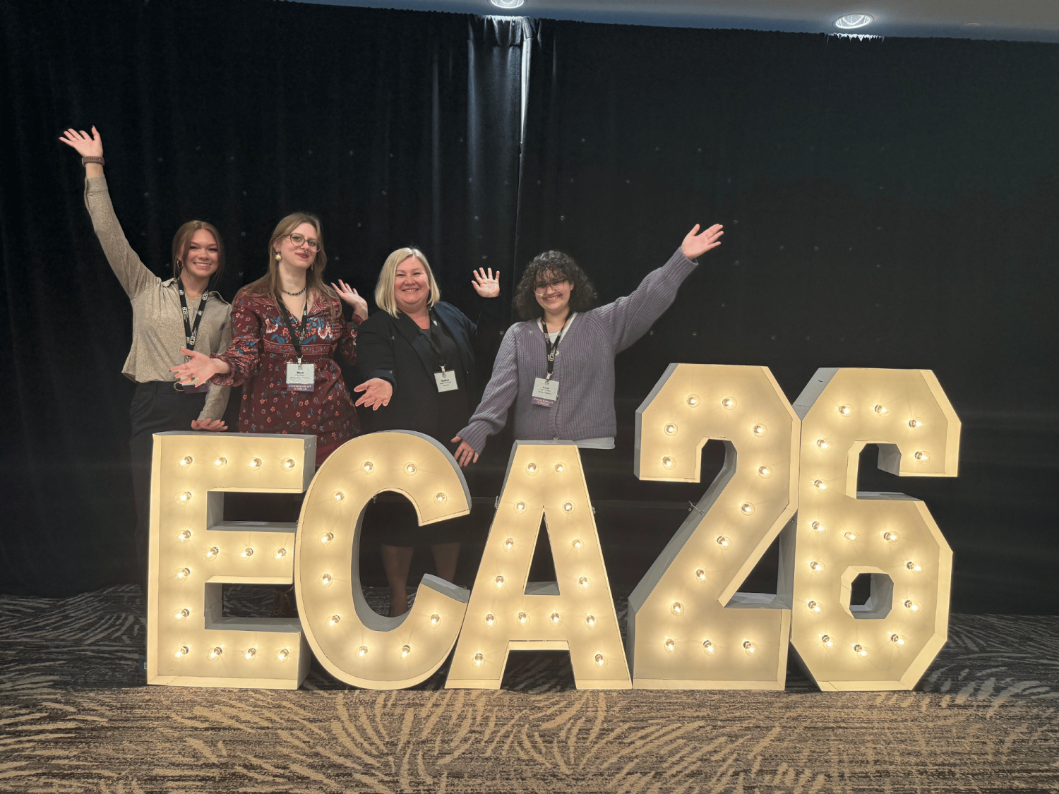 Students and faculty member with their arms outstretched near a large floor sign, "ECA 2026," the conference the group attended