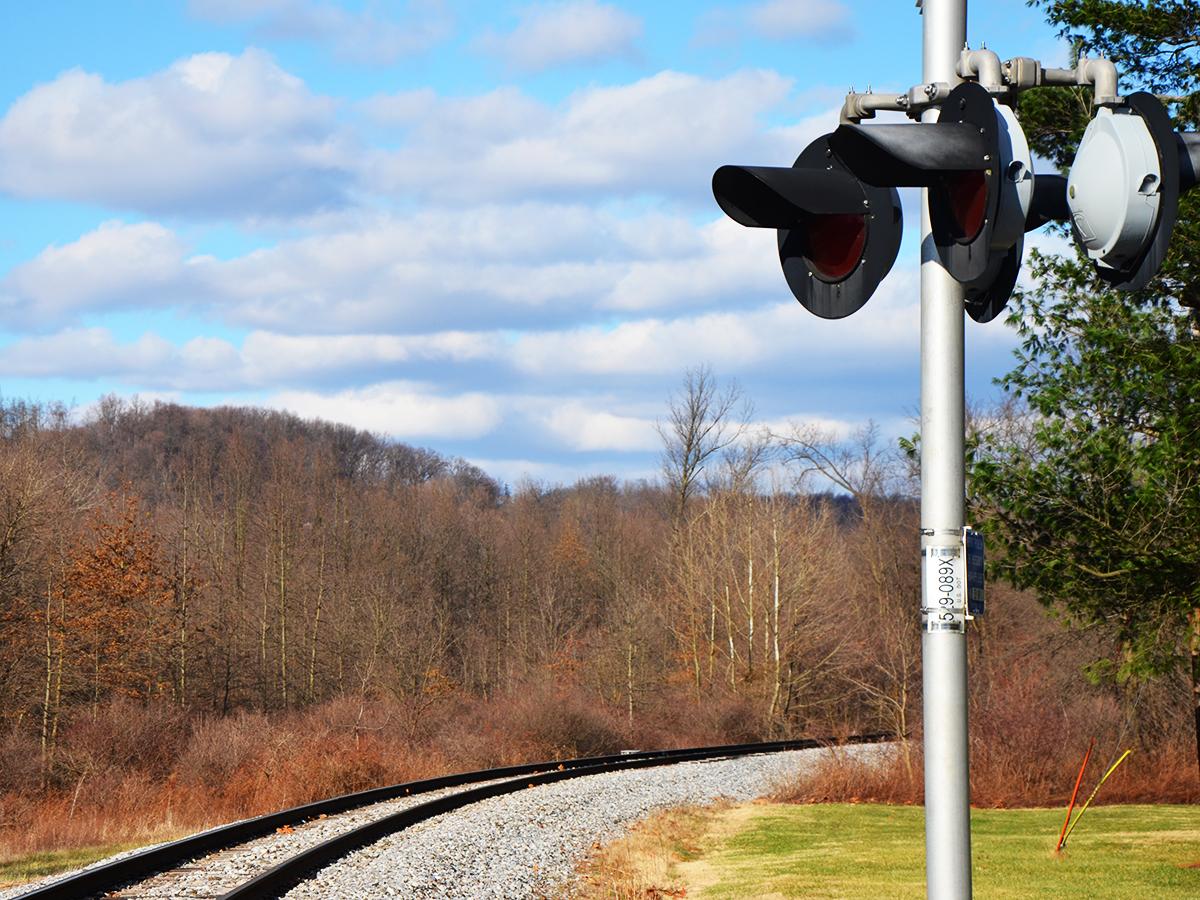 Railroad crossing signal stands beside curving tracks under a blue sky with clouds and leafless trees.