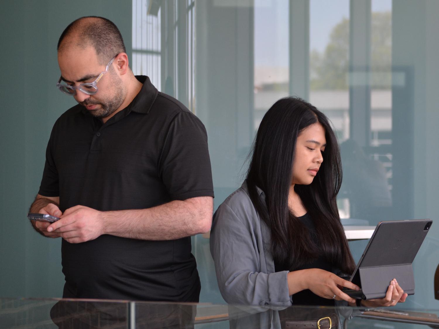 Nelson Roque is looking at a smartphone, and Rinanda Shaleha is looking at a tablet in front of a glass wall