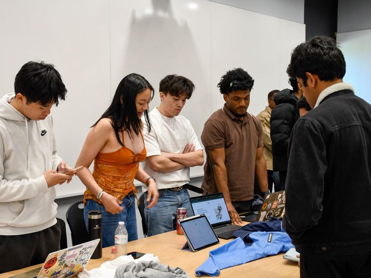 Three people standing in front of a laptop demonstrate their app to several other people in a college classroom. 
