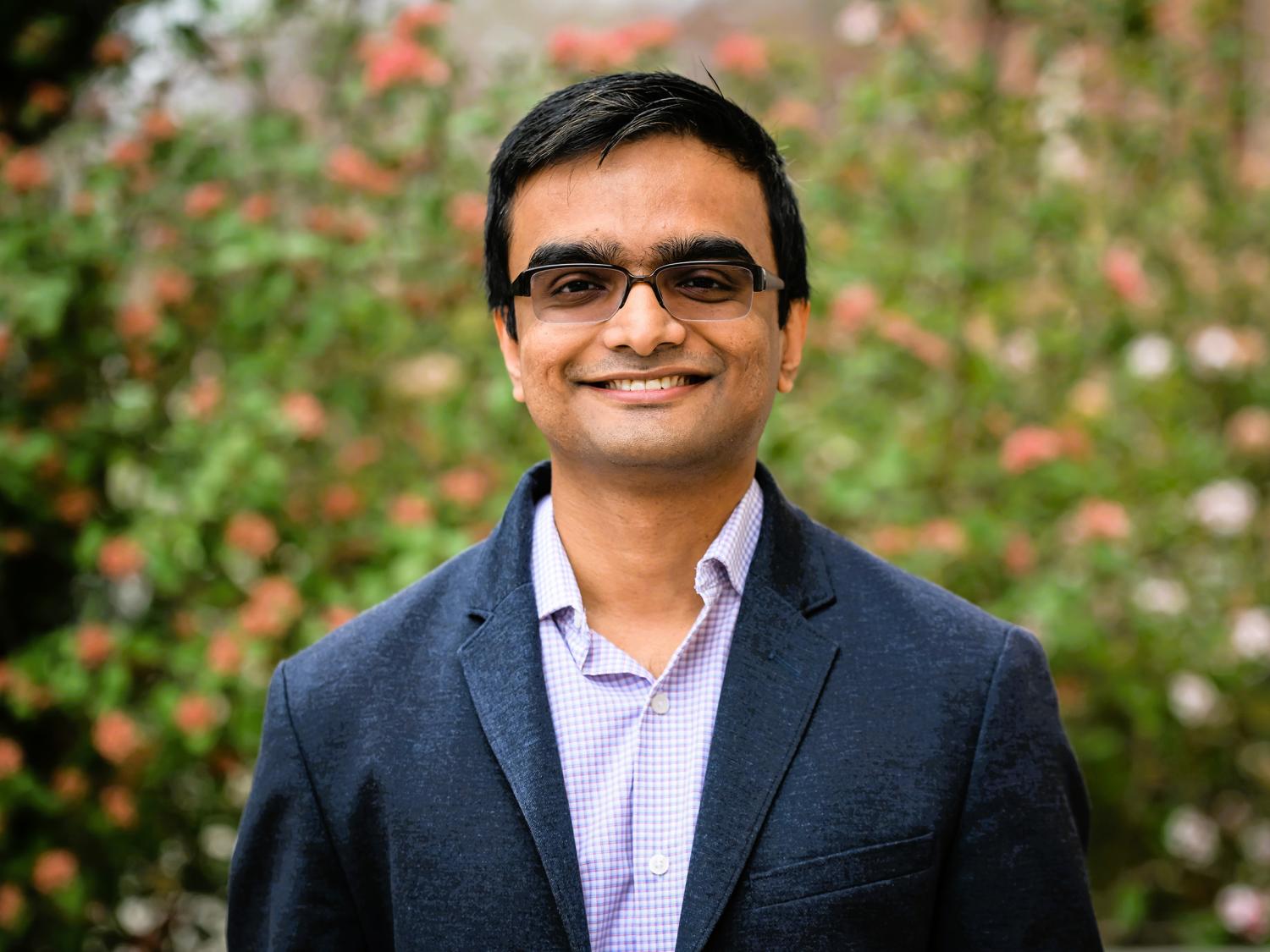 Abhronil Sengupta smiles in a portrait standing in front of a flower bush.