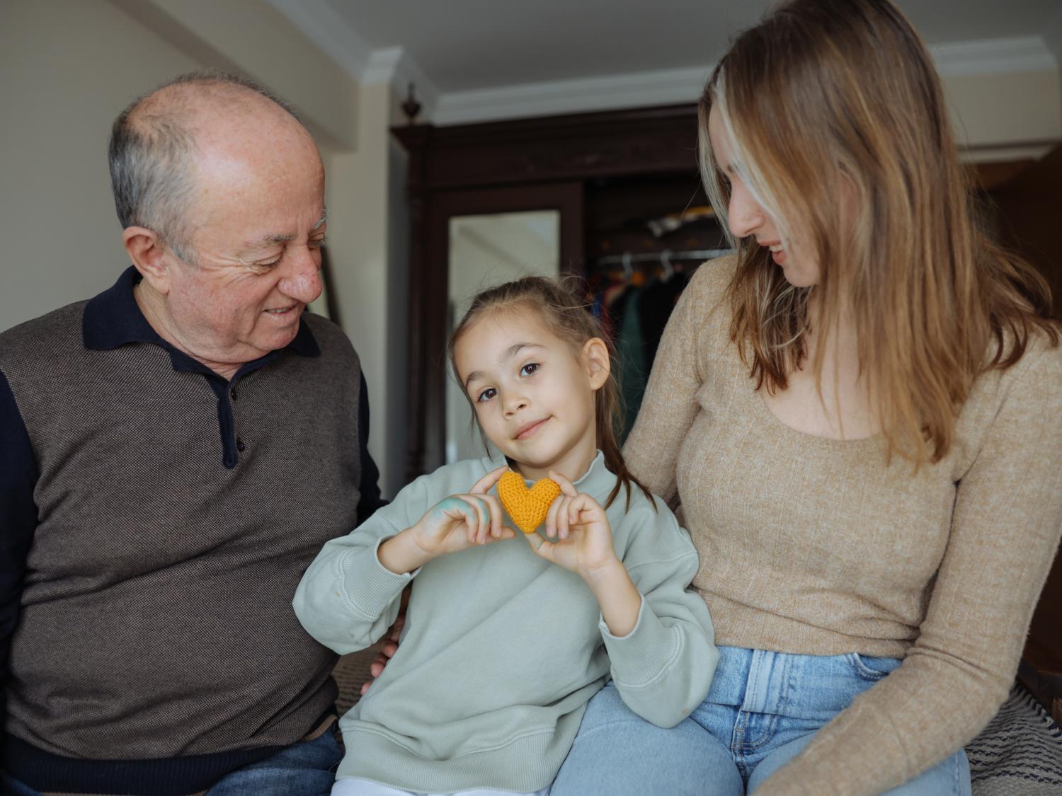Grandfather, daughter and granddaughter in a bedroom. The granddaughter, who is around 6 years old, holds an orange crocheted heart.