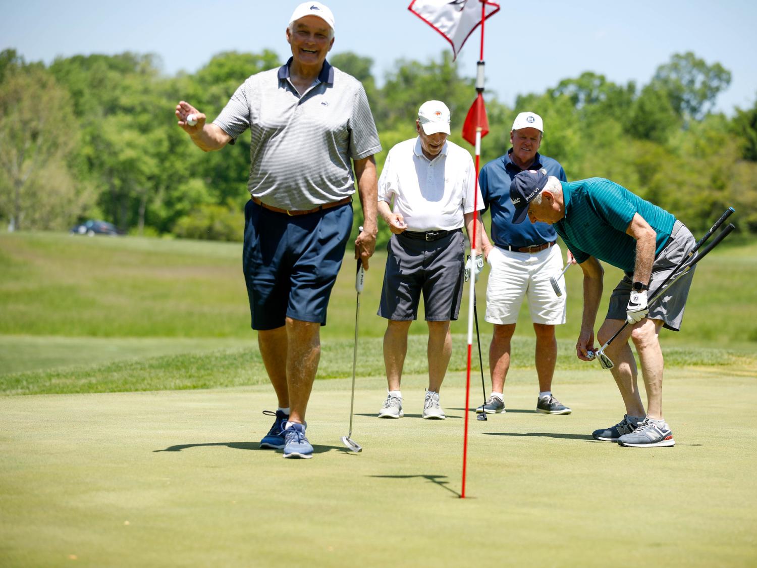 A foursome of golfers gathers around a hole on the golf course, holding clubs and golf balls.