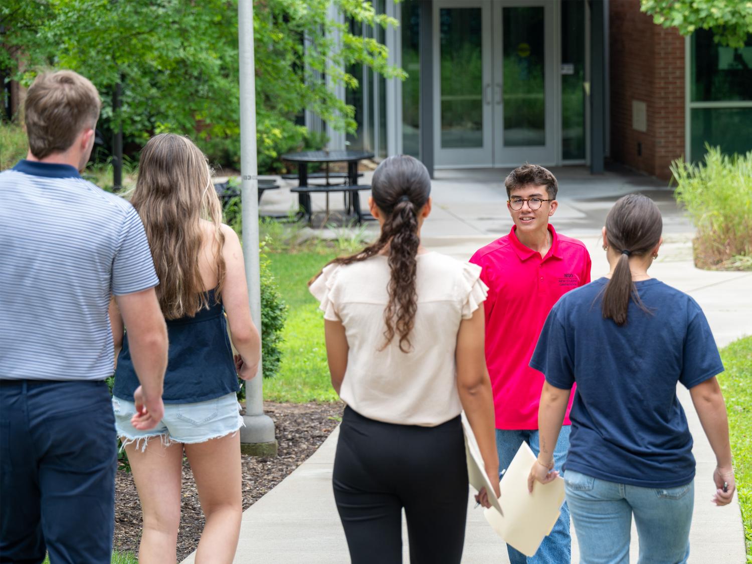 Five people walk along a campus sidewalk outside an academic building. One person holding papers faces the group as they approach, with trees, grass, and outdoor seating visible nearby.