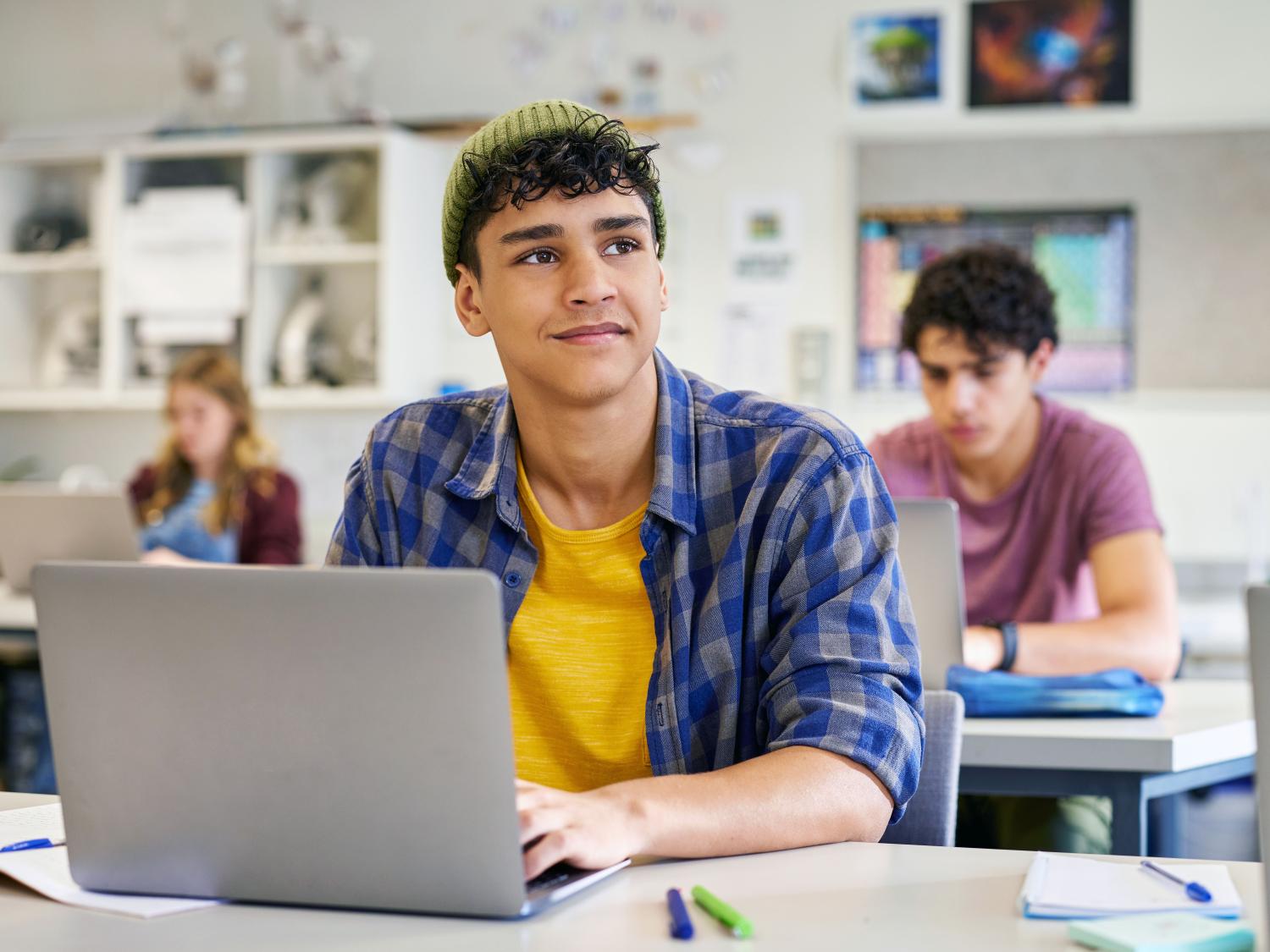 A high school student sits at a laptop in a classroom while looking thoughtfully into the distance, with classmates working in the background. The image illustrates new Penn State research examining how adolescents' career expectations develop and how family socioeconomic background influences the occupations they aspire to pursue.