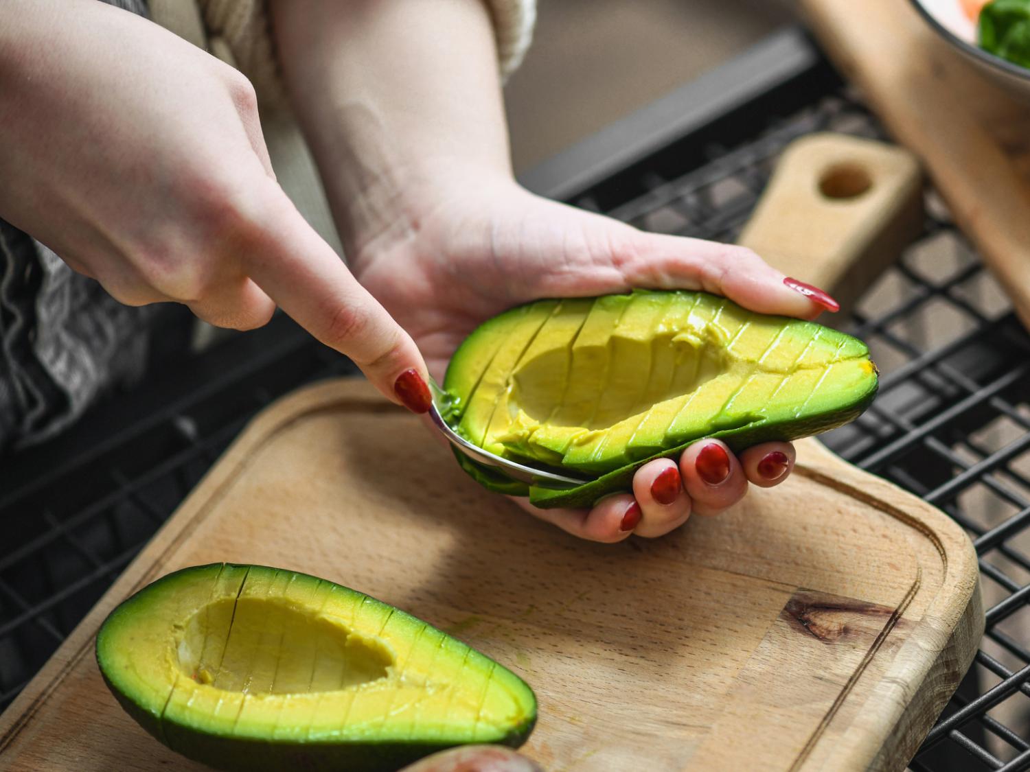 A woman's hands scoop a ripe avocado above a cutting board in a kitchen