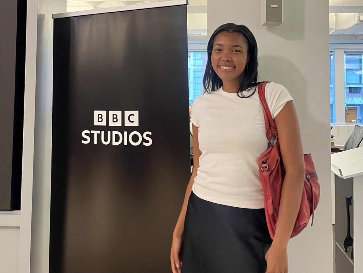 A young woman in a white shirt and black skirt smiles for the camera while standing by a sign that reads "BBC Studios." 