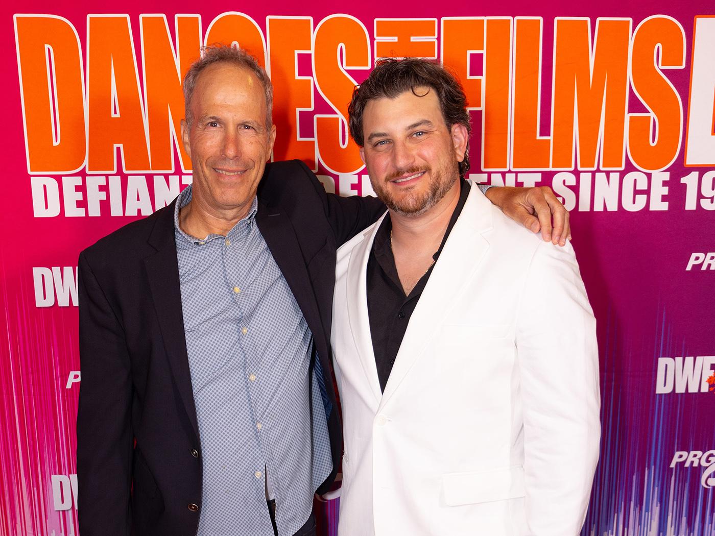 A man in a dark coat and shirt with his arm around a dark-haired man in a white coat.